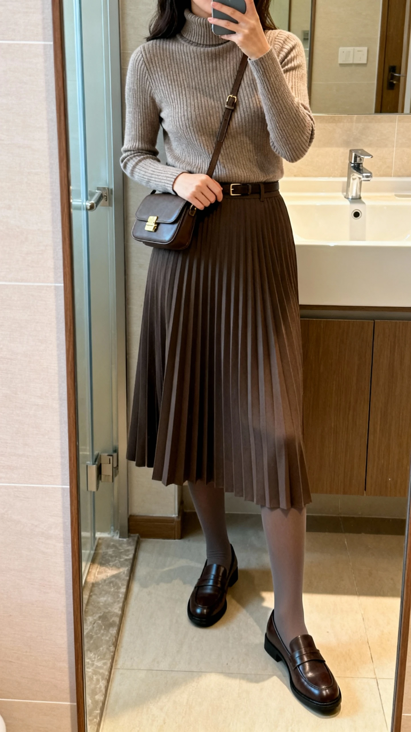 iPhone side-angle mirror selfie of a woman wearing a ribbed turtleneck tucked into a pleated midi skirt, opaque tights, classic loafers, and a small crossbody bag, face not visible, cropped at shoulders, bathroom full-length mirror, natural indoor lighting, iPhone photo quality.