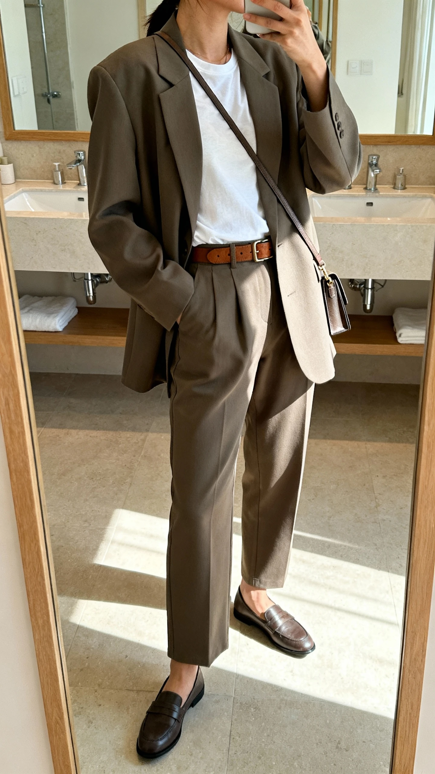 iPhone side-angle mirror selfie of a woman wearing a relaxed tailored suit with an oversized blazer and straight-leg trousers over a simple white tee, paired with loafers, a leather belt, and a slim crossbody bag, face not visible, bathroom mirror, indoor natural light, iPhone photo quality.