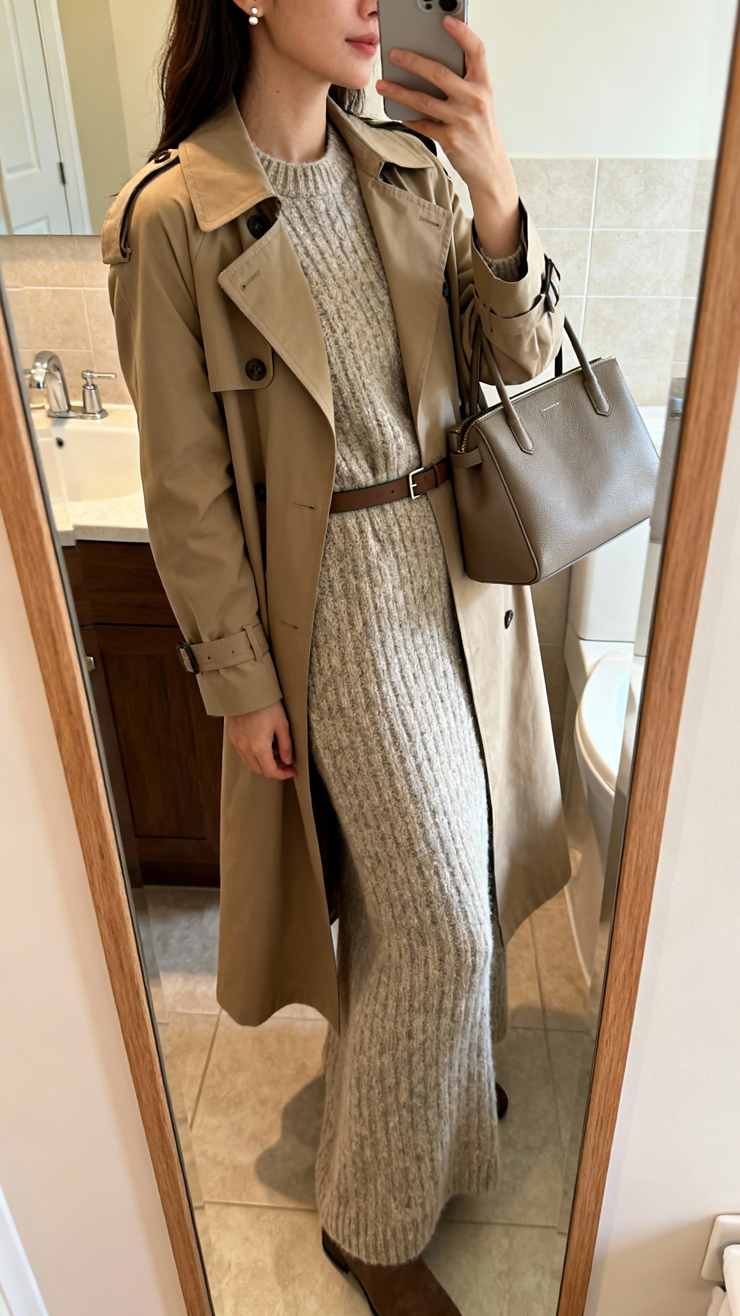 iPhone side-angle mirror selfie of a woman wearing a cozy knit maxi dress under a classic trench coat with a slim belt, subtle stud earrings, and a structured tote, face not visible, bathroom full-length mirror, morning indoor lighting, iPhone photo quality