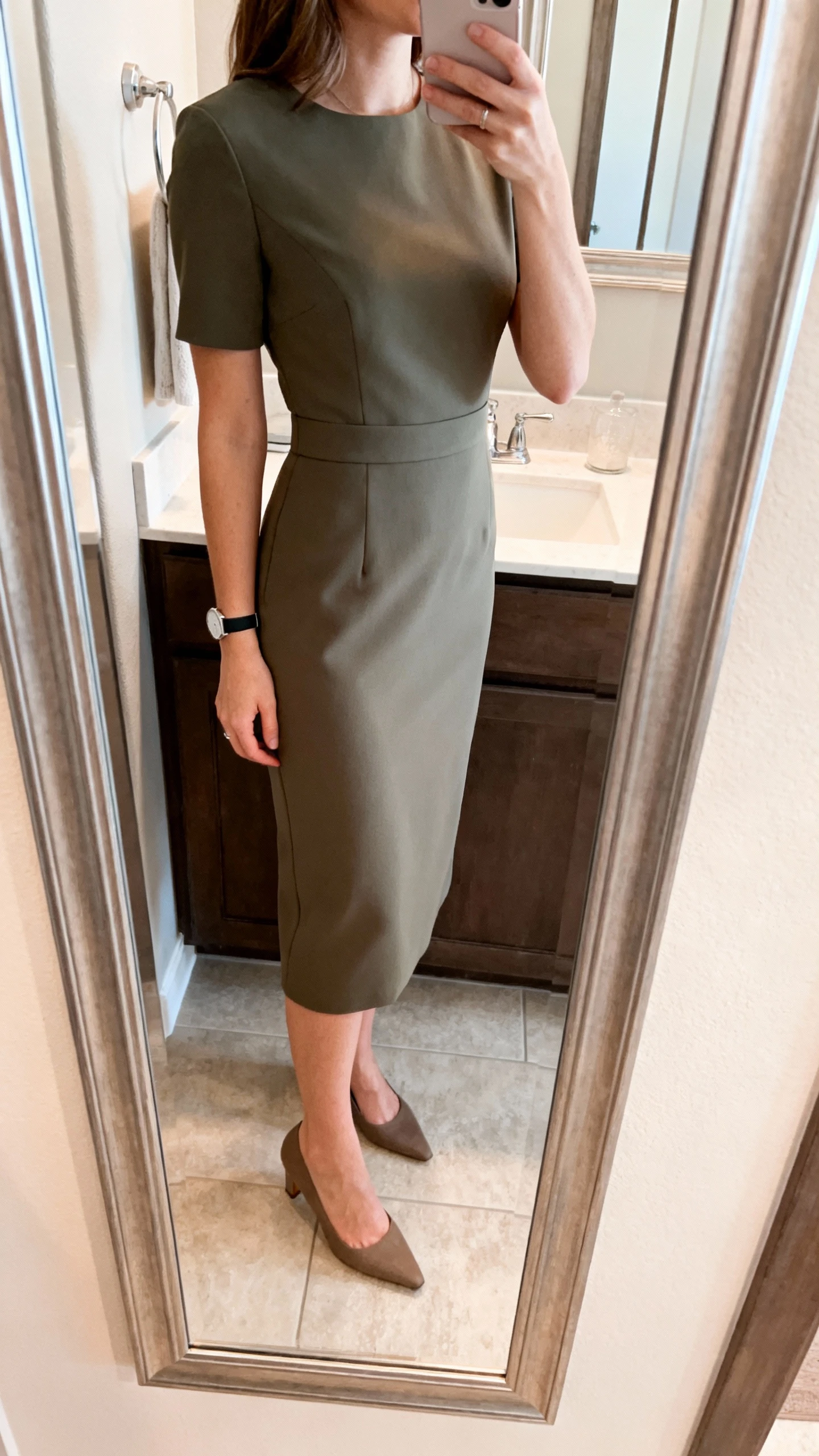 iPhone side-angle mirror selfie of a woman wearing a tailored midi sheath dress with a gentle defined waist and clean lines, paired with classic closed-toe heels and a minimal watch, face not visible, bathroom mirror, soft indoor lighting, iPhone photo quality.