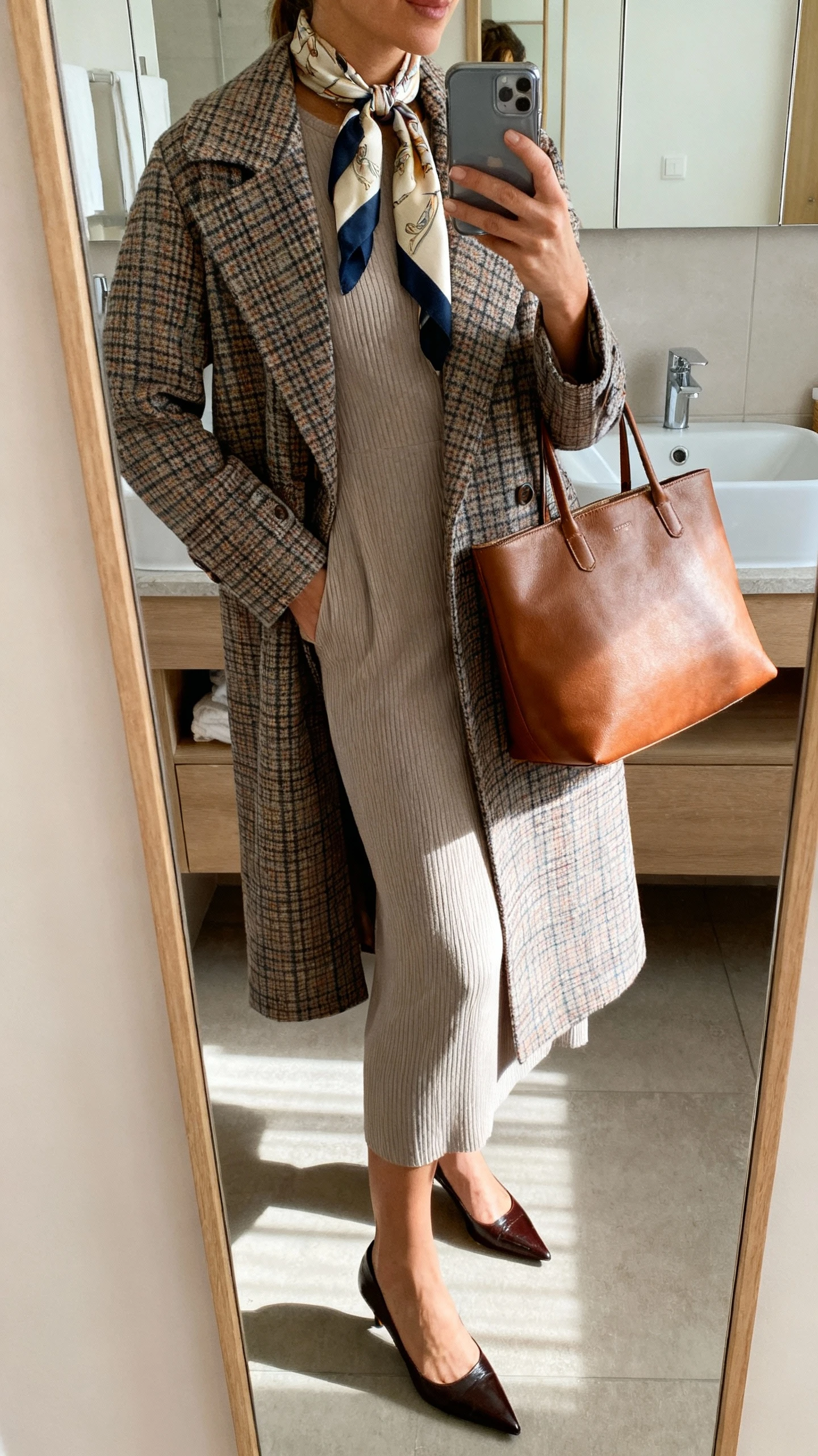 iPhone side-angle mirror selfie of a woman wearing a plaid midi coat layered over a column dress, pointed toe pumps, leather tote, and a silk scarf draped at the neckline, face not visible, bathroom mirror, morning light, iPhone photo quality.