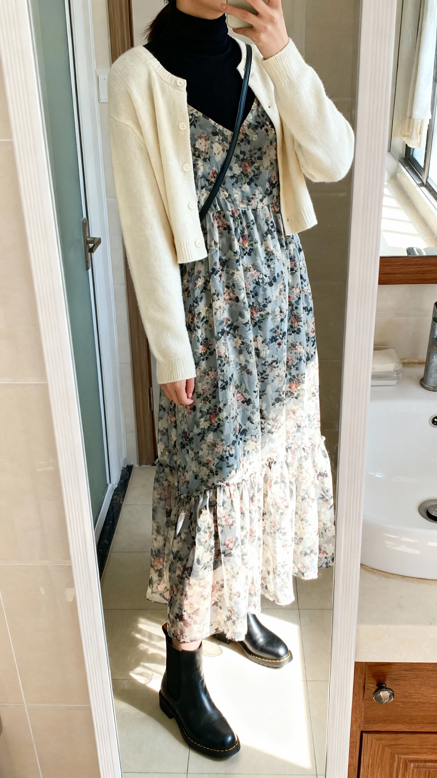 iPhone side-angle mirror selfie of a woman wearing a floral lightweight maxi dress layered over a fitted black turtleneck, with a cropped cream cardigan, black Chelsea boots, and a simple crossbody, face not visible, cropped at shoulders, bathroom mirror, bright natural light, iPhone photo quality.