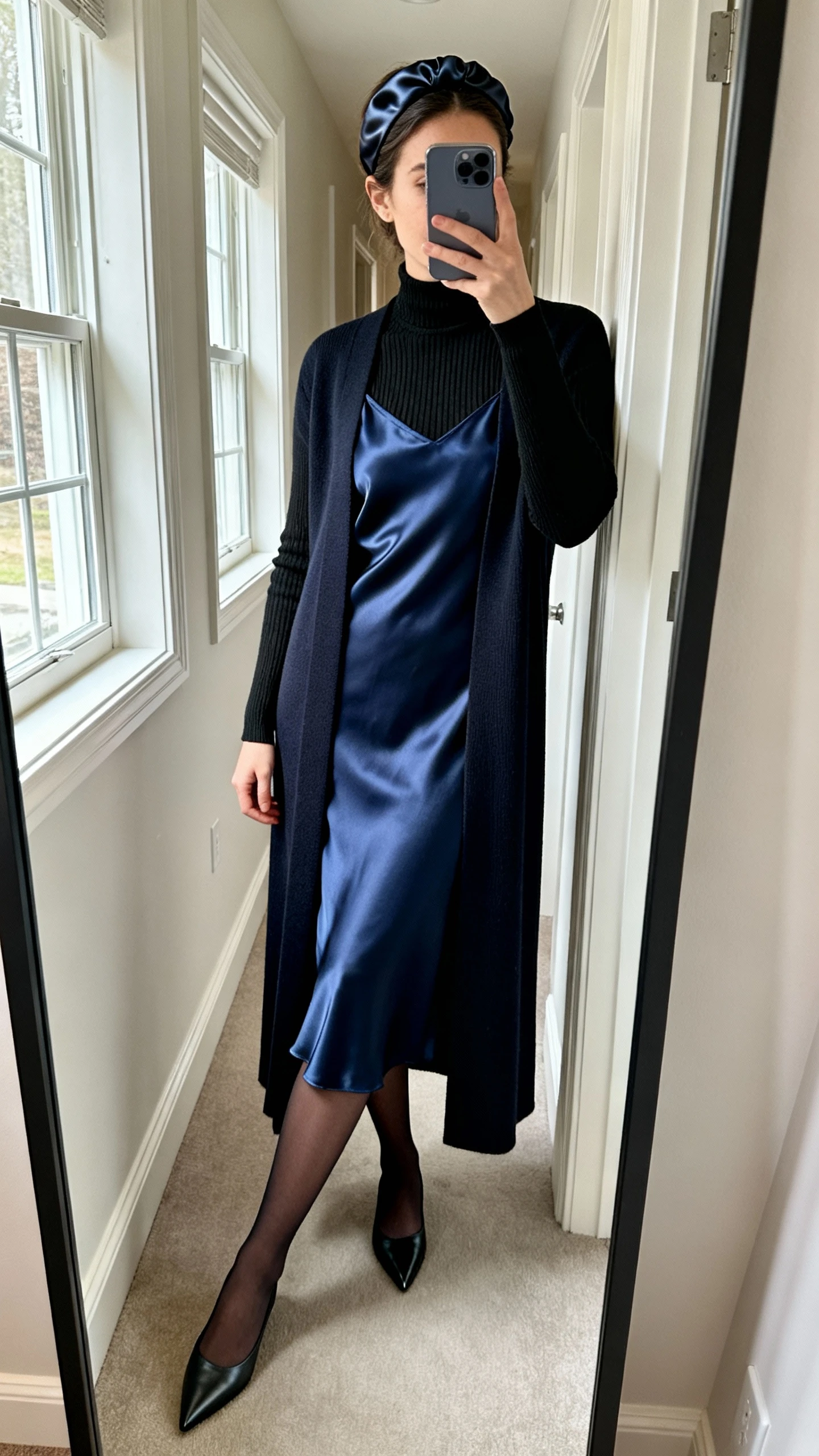 iPhone side-angle mirror selfie of a woman wearing an ink blue midi satin slip dress layered over a fitted black fine-gauge turtleneck, longline tailored cardigan, pointed-toe flats, matte opaque tights, and a satin headband; face not visible, cropped at shoulders, full-length mirror in hallway, soft window light, iPhone photo quality.