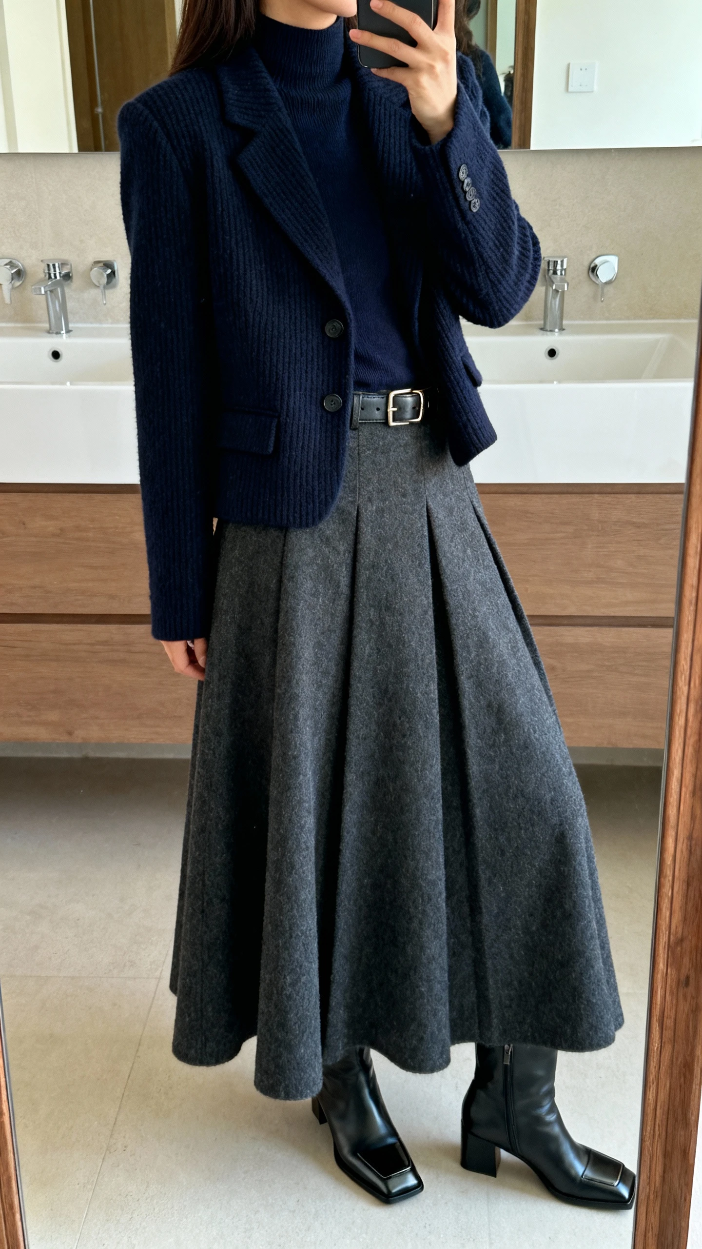 iPhone side-angle mirror selfie of a woman wearing a tailored navy sweater blazer, full A-line maxi skirt in wool blend, refined mock-neck top, classic leather belt, and square-toe heeled boots, face not visible, cropped at shoulders, bathroom mirror with soft indoor lighting, iPhone photo quality.