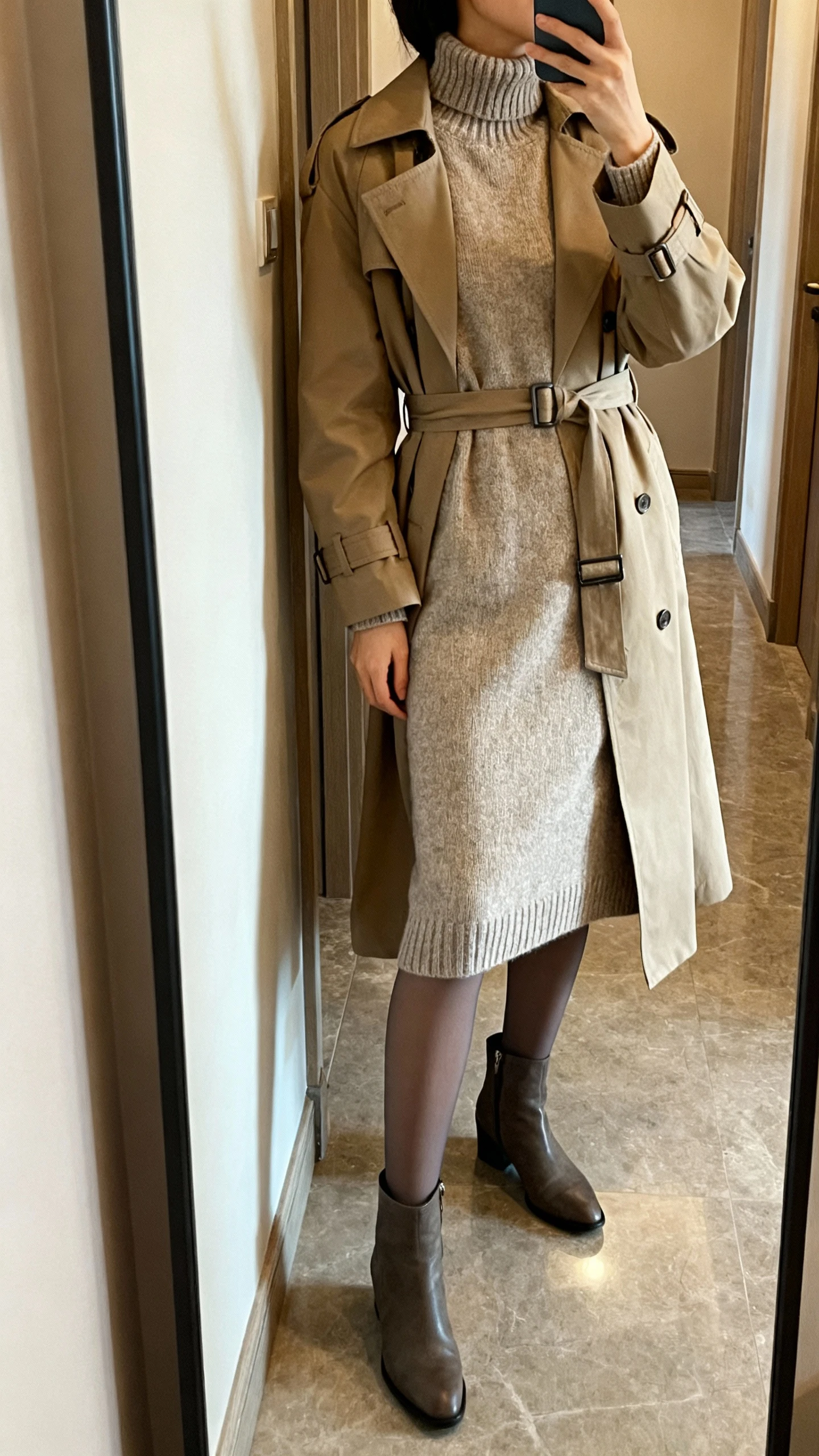 iPhone side-angle mirror selfie of a woman wearing a knit mock-neck midi dress layered with a belted trench coat, opaque tights, and ankle boots, face not visible, hallway mirror, soft indoor lighting, iPhone photo quality
