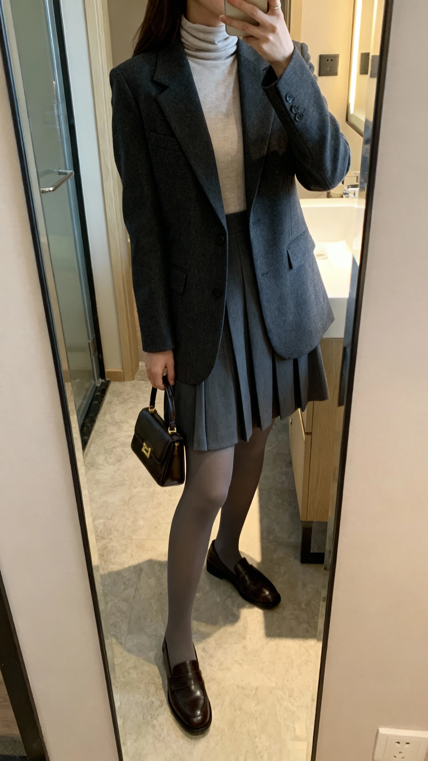 iPhone side-angle mirror selfie of a woman wearing a high-neck A-line dress layered with a tailored blazer, opaque tights, and classic loafers, carrying a small structured handbag, face not visible, cropped at shoulders, bathroom mirror, indoor lighting, iPhone photo quality.
