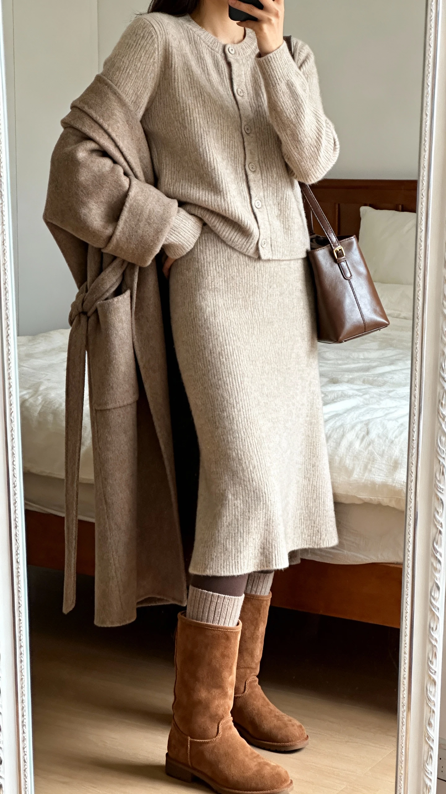 iPhone side-angle mirror selfie of a woman wearing a matching knit set (long-sleeve cardigan and A-line midi skirt) with thermal leggings underneath, tall suede boots, an oversized wrap coat, and a medium leather shoulder bag, face not visible, cropped at shoulders, bedroom mirror with natural daylight, iPhone photo quality.