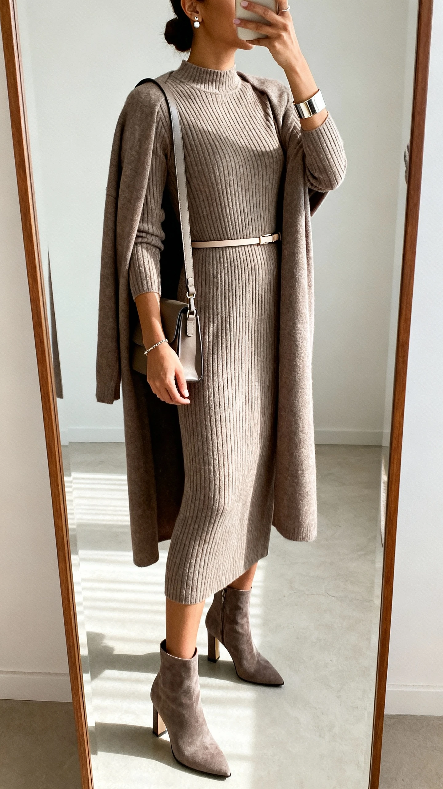 iPhone side-angle mirror selfie of a woman wearing a knit midi column dress with a higher neckline and long sleeves, thin waist belt, heeled ankle boots, structured shoulder bag, stud earrings, and a cuff bracelet; longline cardigan over shoulders, face not visible, full-length mirror, soft indoor lighting, iPhone photo quality.