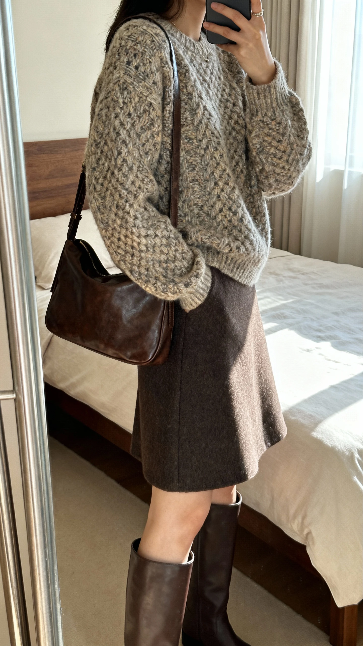 iPhone side-angle mirror selfie of a woman wearing a textured knit sweater and a wool A-line skirt, with knee-high boots and a leather shoulder bag, face not visible, bedroom mirror, soft afternoon window light, iPhone photo quality.
