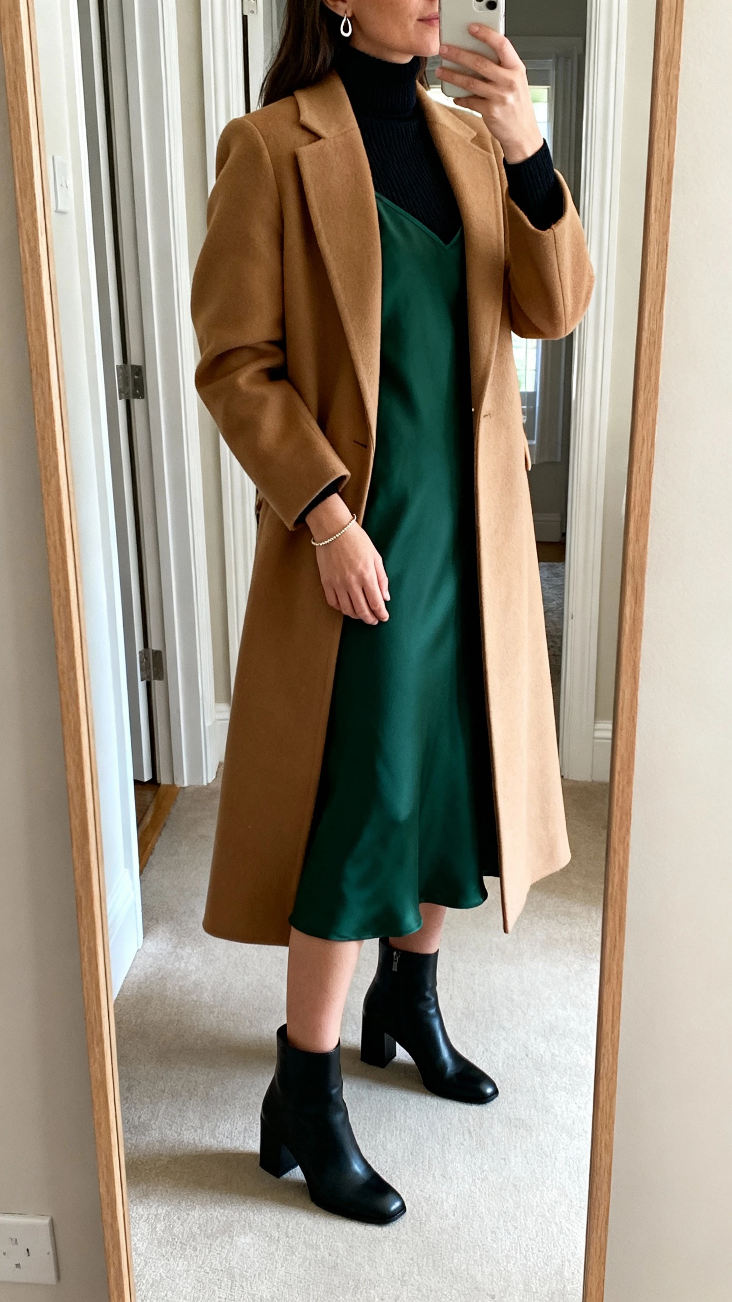 iPhone side-angle mirror selfie of a woman wearing a deep green midi slip dress over a fitted black merino turtleneck, long tailored camel coat, black block-heel ankle boots, simple drop earrings and a delicate bracelet, face not visible, full-length mirror in entryway, soft morning light, iPhone photo quality.