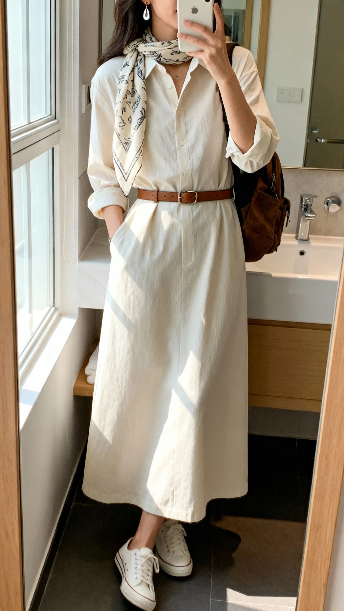 iPhone side-angle mirror selfie of a woman wearing a maxi shirt dress with three-quarter sleeves and a modest collar, a slim leather belt, minimal white sneakers, a lightweight printed scarf, simple drop earrings, and a small backpack, face not visible, bathroom mirror, natural window light, iPhone photo quality.
