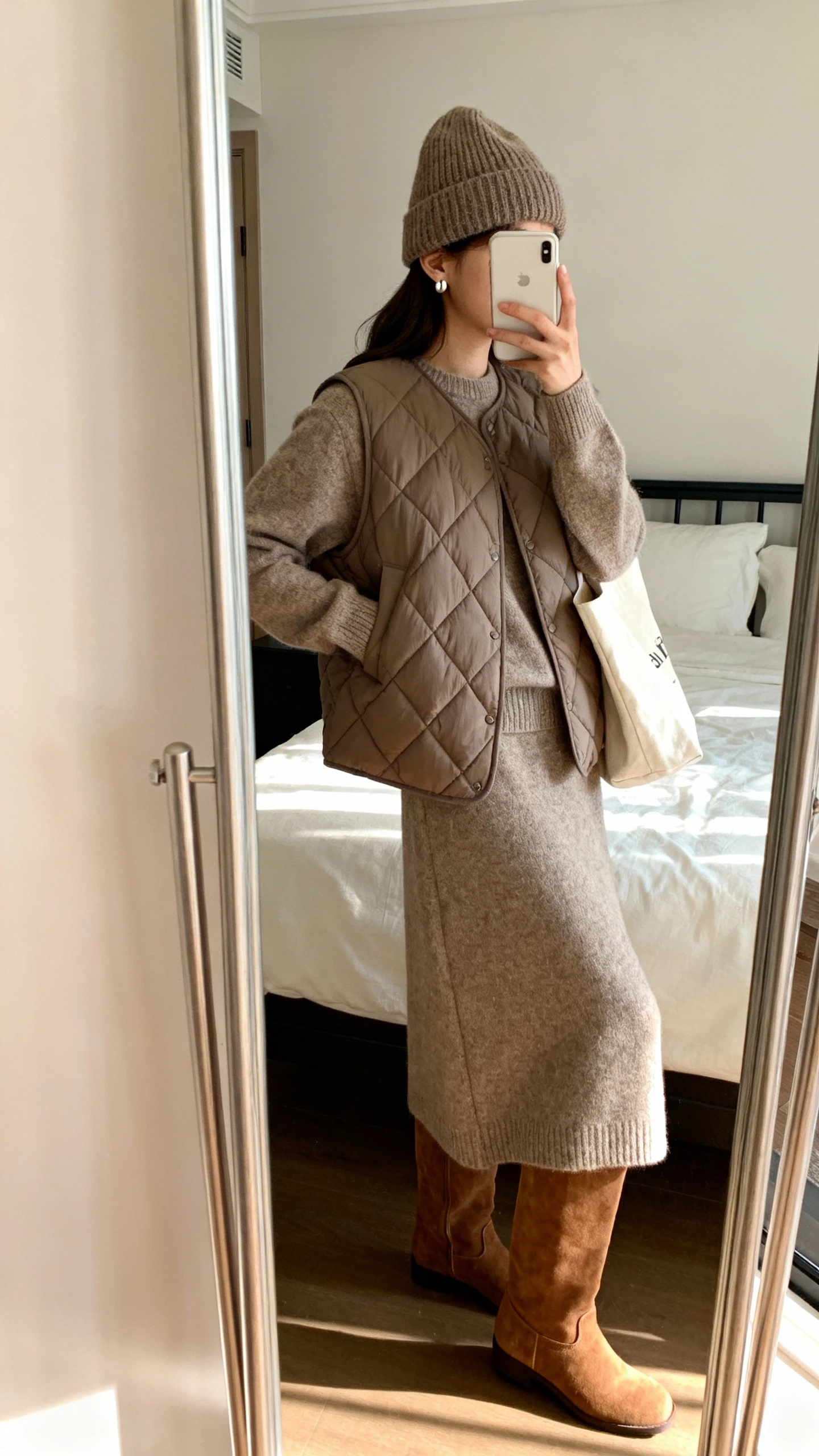 iPhone side-angle mirror selfie of a woman wearing a sweater dress with a quilted vest layered over and the dress skirt styled as a long skirt silhouette, tall suede boots, knit beanie, simple stud earrings, tote bag, face not visible, cropped at neck, bedroom mirror, natural afternoon light, iPhone photo quality.