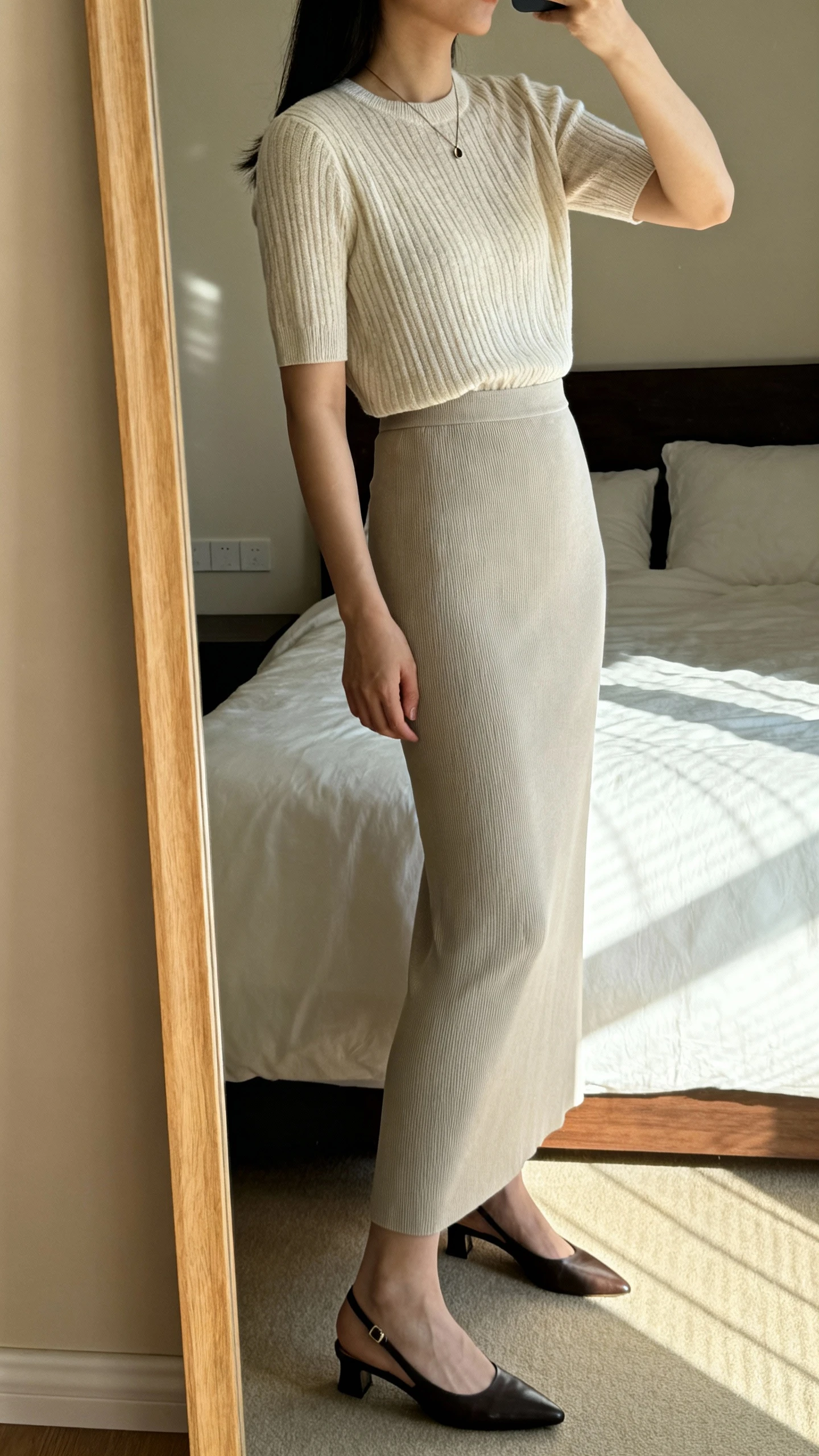 iPhone side-angle mirror selfie of a woman wearing a fine-gauge knit top tucked into a long column skirt, with sleek slingback flats and a minimal pendant, face not visible, full-length mirror in bedroom, soft morning light, iPhone photo quality.