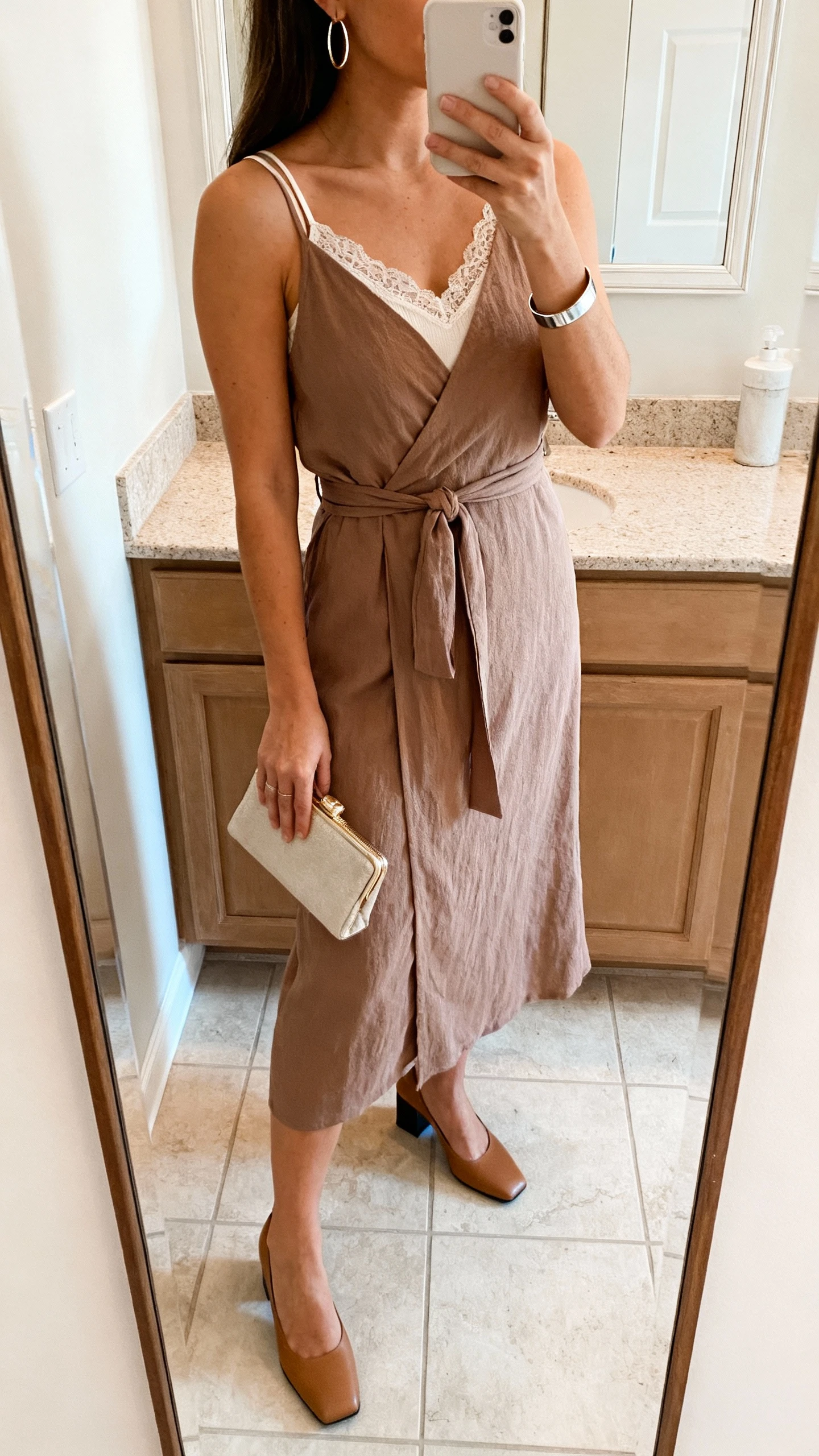 iPhone side-angle mirror selfie of a woman wearing a midi wrap dress with a secure tie and modest V-neck, low block heels, a thin lace-trim layering tank for extra coverage, a compact clutch, simple hoop earrings, and a sleek cuff bracelet, face not visible, bathroom mirror, soft natural light, iPhone photo quality.