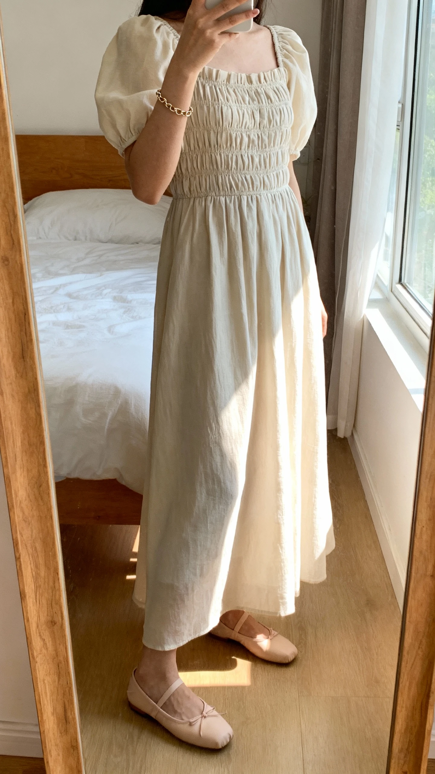 iPhone side-angle mirror selfie of a woman wearing a smocked-bodice maxi dress with soft puff sleeves, ballet flats, and a delicate chain bracelet, face not visible, bedroom mirror, afternoon window light, iPhone photo quality.