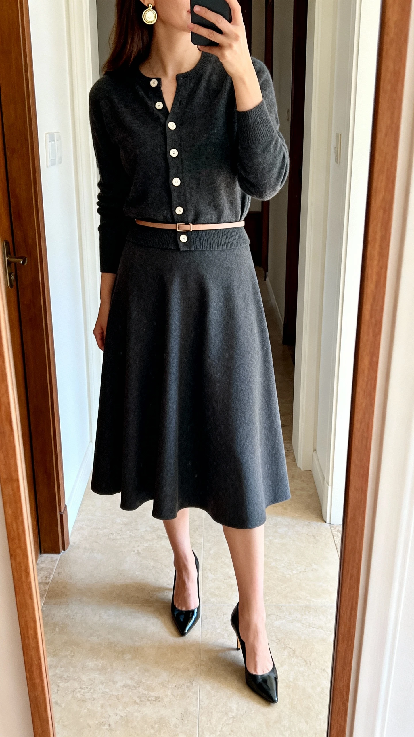 iPhone side view mirror selfie of a woman in an A-line midi skirt with a buttoned-up cardigan, thin waist belt, kitten heels, vintage-inspired clip-on earrings, face not visible, cropped at shoulders, entryway mirror, natural daylight, iPhone photo quality.
