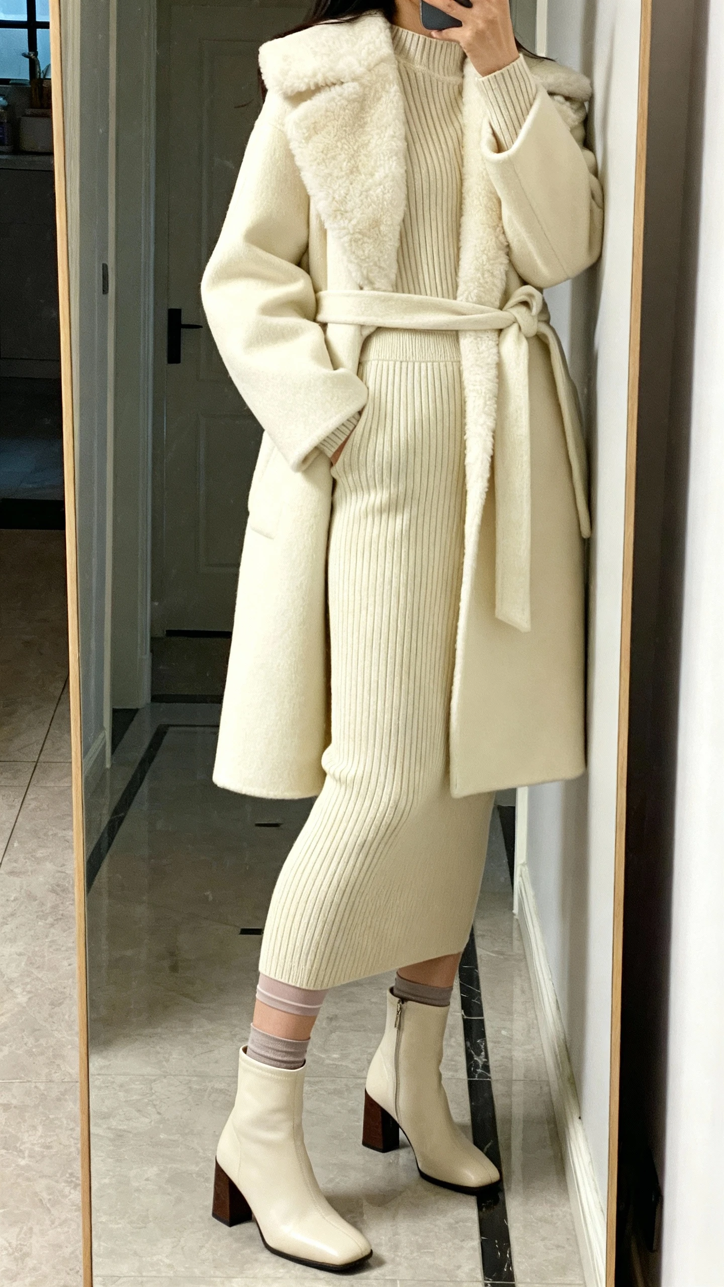 iPhone side-view mirror selfie of a woman wearing a polished knit set (pencil skirt and matching sweater), a cream shearling-lined wrap coat, block-heel boots, and a thermal slip under the skirt for smooth lines, face not visible, full-length mirror in entryway with indoor lighting, iPhone photo quality.
