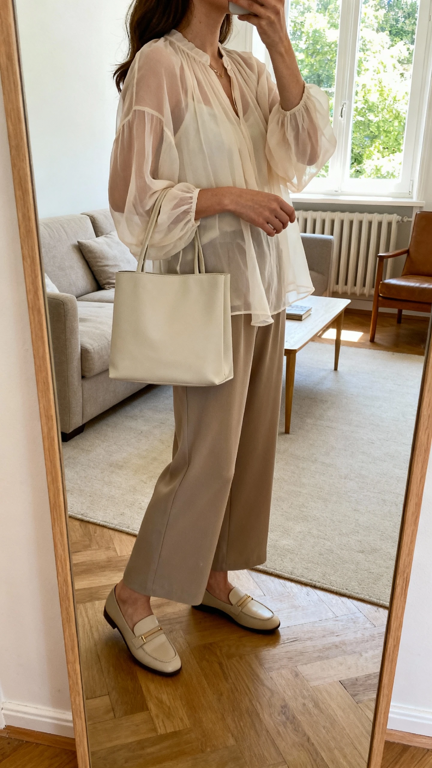 Casual iPhone mirror selfie of a woman wearing a floaty chiffon blouse with wide-leg pants, loafers, and a minimalist tote, face not visible, side angle in living room mirror with natural window light, iPhone photo quality.