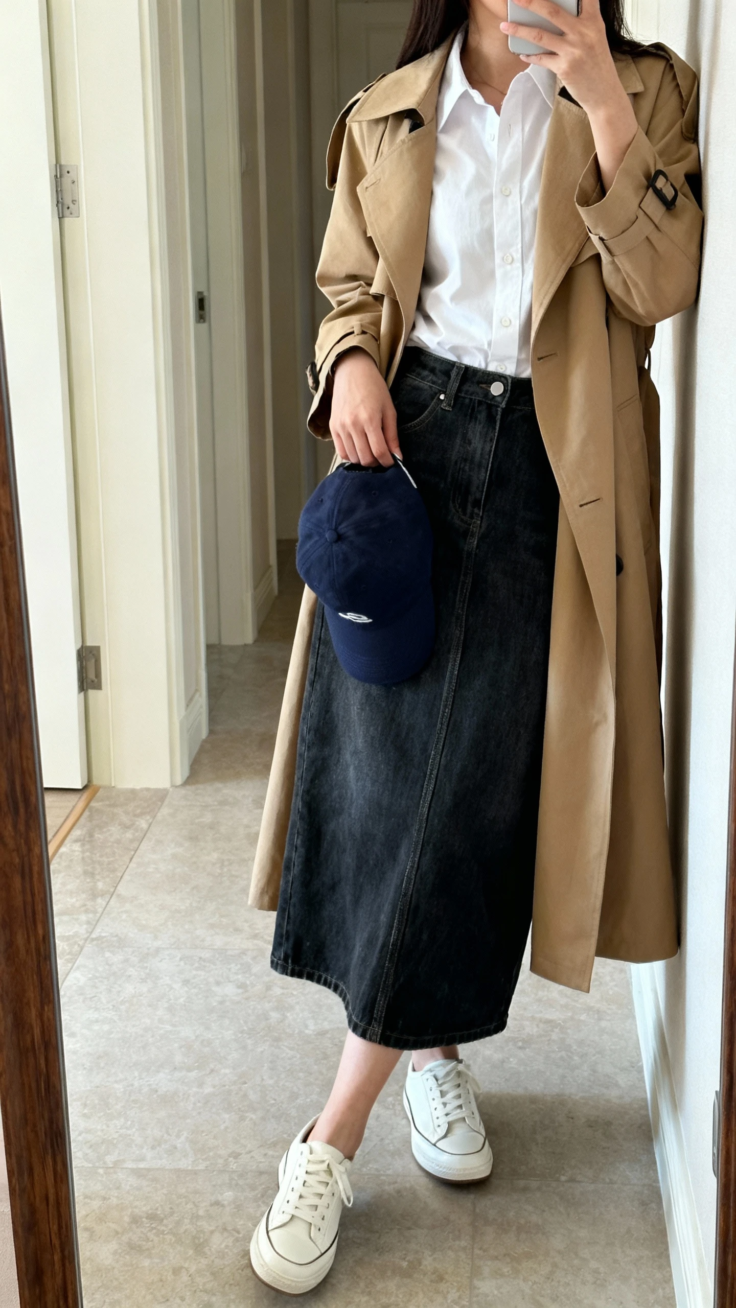 Casual iPhone side-angle mirror selfie of a woman in a dark-wash denim maxi skirt, a crisp white button-up shirt, a lightweight tan trench, clean white leather sneakers, and a navy baseball cap in hand; face not visible, full-length hallway mirror, natural daylight, iPhone photo quality.