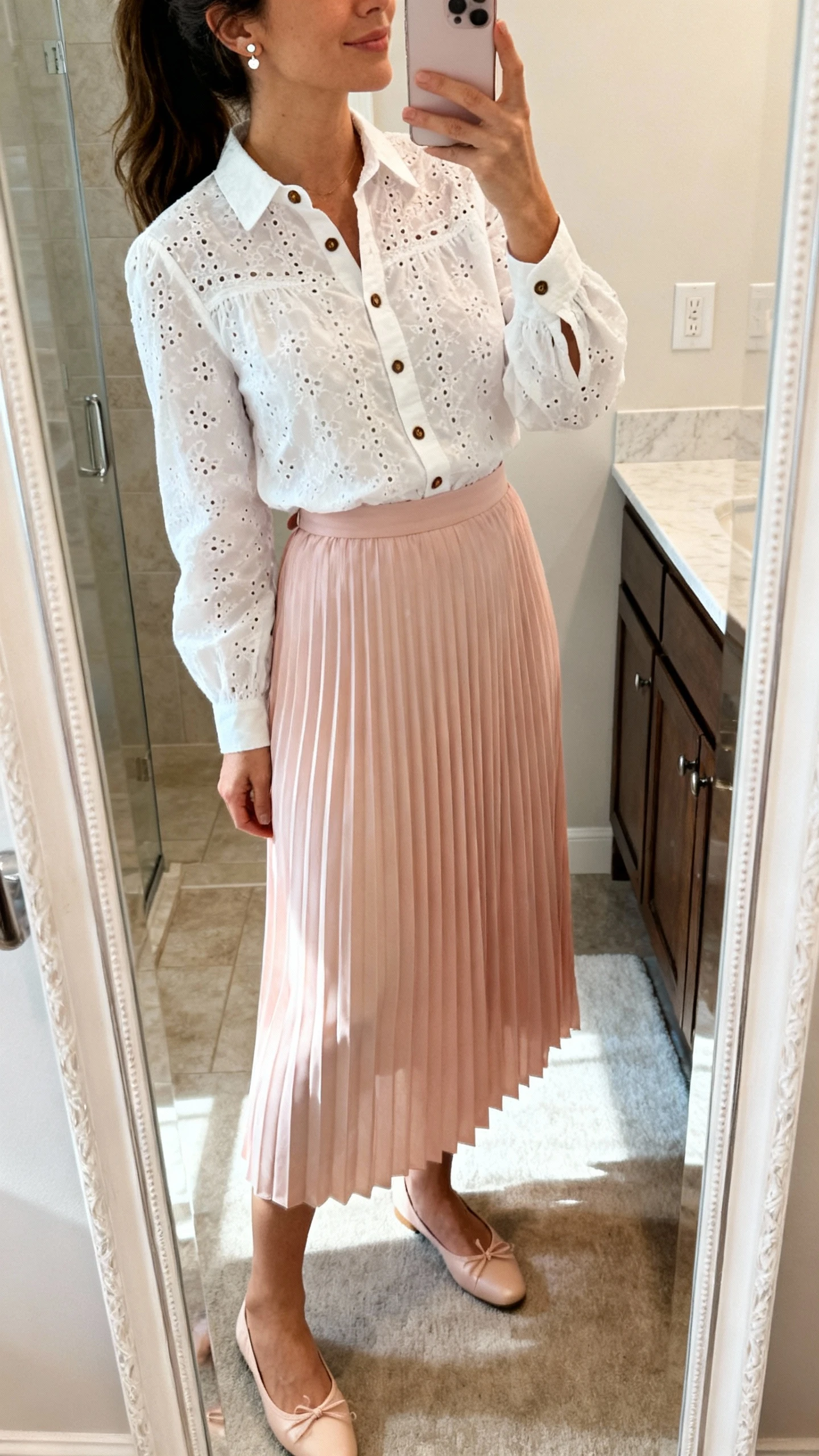 iPhone back/side mirror selfie of a woman wearing a buttoned-up white eyelet blouse tucked into a pastel pleated midi skirt, with dainty stud earrings and ballet flats; face not visible, bathroom mirror, morning indoor lighting, iPhone photo quality.