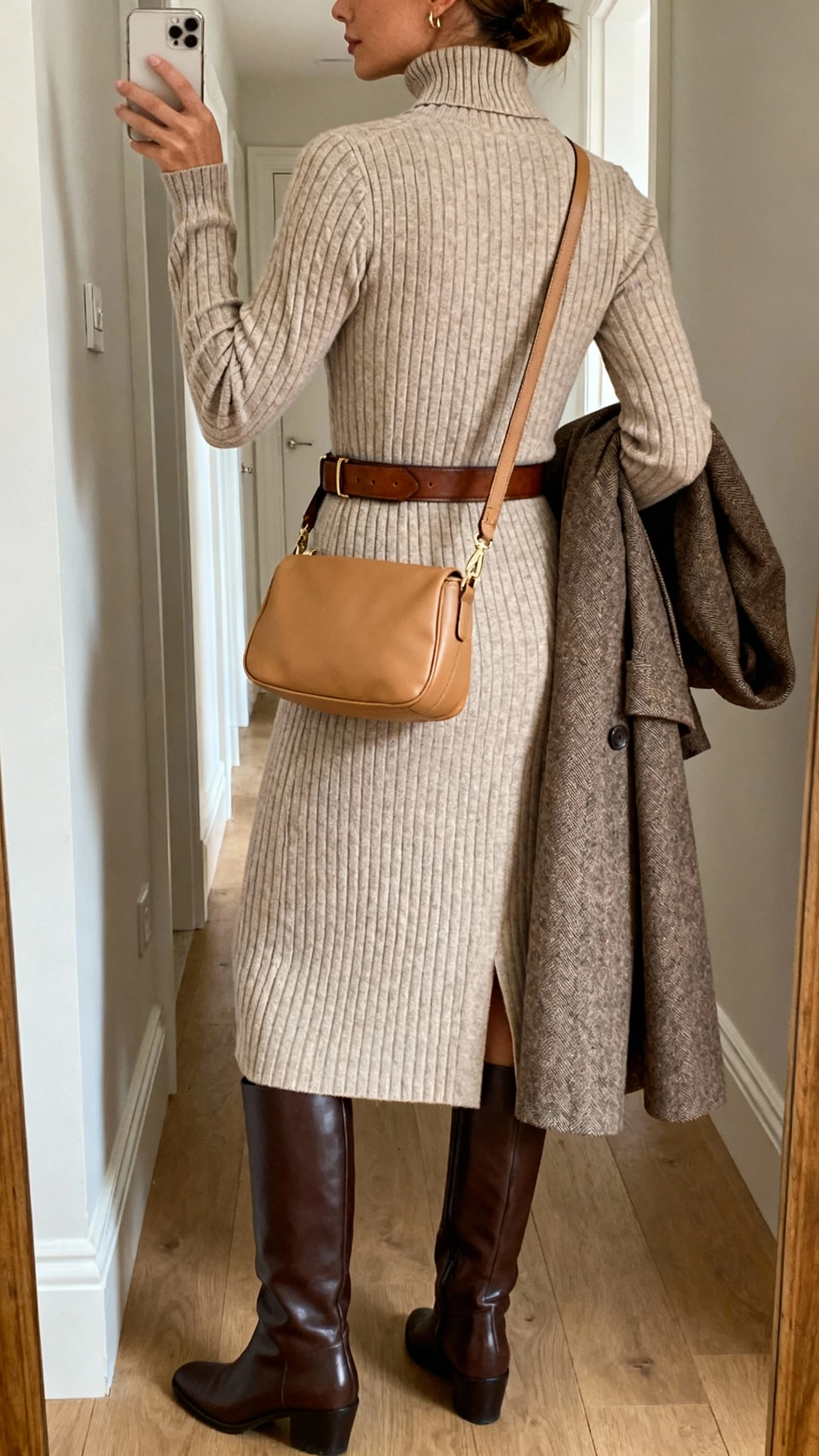iPhone back-view hallway mirror selfie of a woman wearing a ribbed knit midi dress with long sleeves and a mock neck, leather knee-high boots, a classic leather belt, a tailored wool coat carried over one arm, and a medium camel crossbody bag; face not visible, natural indoor lighting, iPhone photo quality.