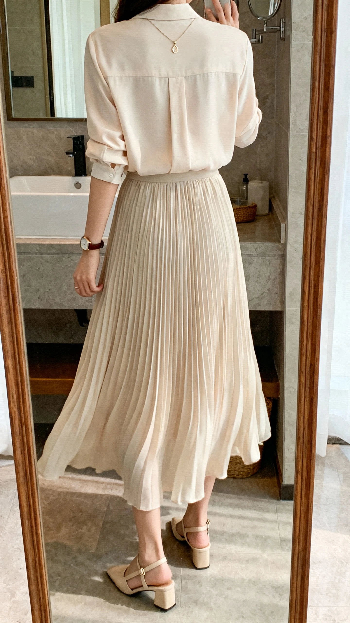 iPhone back-view mirror selfie of a woman wearing a flowing pleated skirt with a soft blouse tucked in, delicate pendant necklace, slingback heels, and a slim leather watch; face not visible, bathroom mirror with soft indoor lighting, casual iPhone photo.