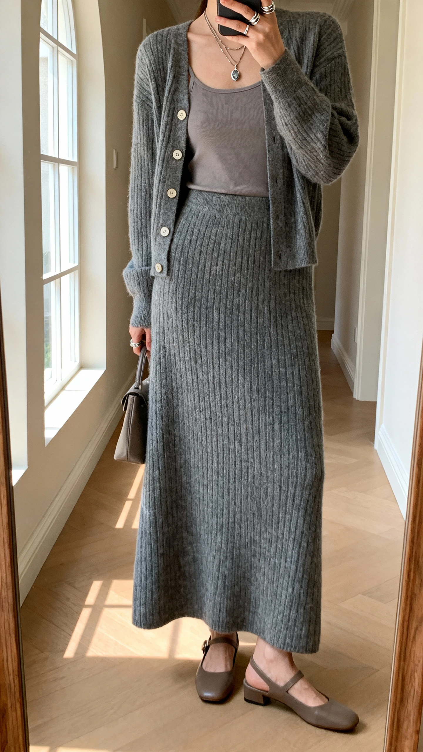 iPhone back-view mirror selfie of a woman wearing a slate knit maxi skirt with a matching buttoned cardigan over a simple camisole, low slingback flats, layered pendant necklace, and stacked rings with a small bag in hand, face not visible, hallway full-length mirror, soft window light, iPhone photo quality.