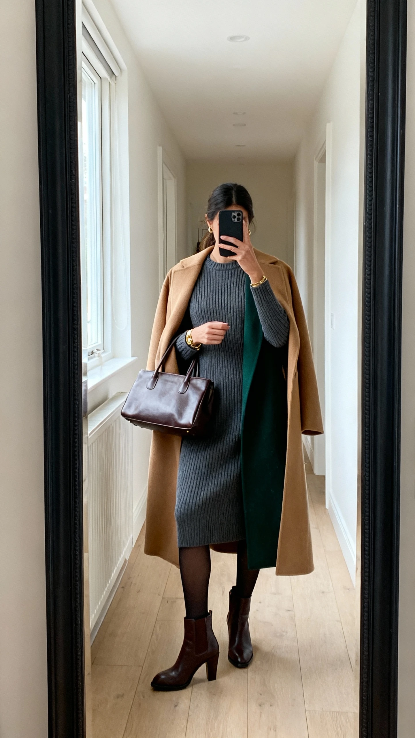 iPhone back-view mirror selfie of a woman wearing a knit midi dress with a crew neck and long sleeves (ribbed or smooth), a longline coat in camel/charcoal/deep green, heeled ankle boots or classic Chelsea boots, opaque tights in black or dark brown, a structured leather tote, and minimal gold or silver jewelry; face not visible, hallway full-length mirror, morning window light, iPhone photo quality.