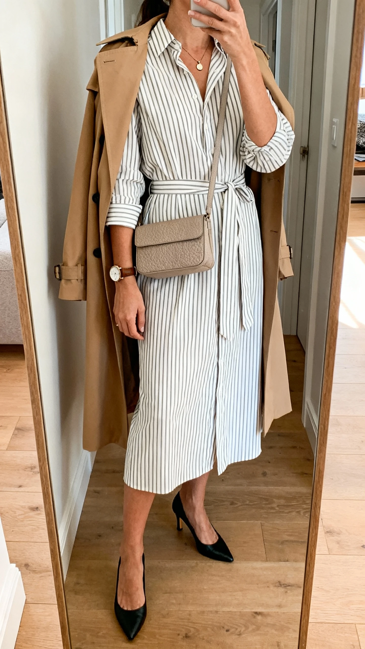 iPhone back-view mirror selfie of a woman wearing a vertical-striped midi shirt dress with a tie belt, refined black kitten-heel slingbacks, camel trench coat draped over shoulders, small pendant necklace, leather-strap watch, and a textured neutral leather crossbody; face not visible, hallway full-length mirror, indoor daylight, iPhone photo quality.