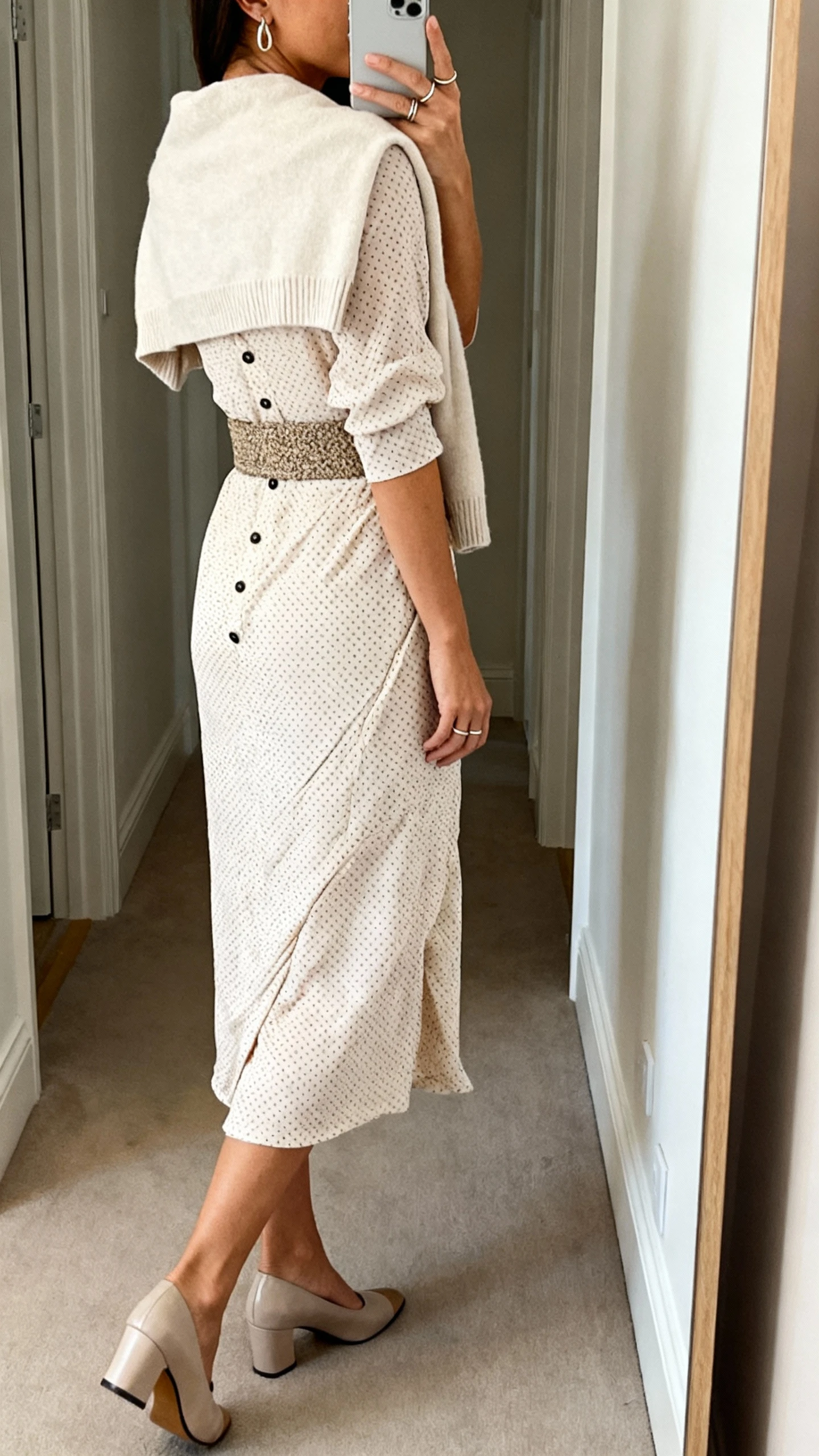 iPhone back-view mirror selfie of a woman wearing an opaque Swiss dot faux-wrap midi dress with a snap at the bust, soft cardigan or cropped knit shrug, textured belt for definition, closed-toe slingback heels or dressy loafers, minimal drop earrings and slim stacking rings; face not visible, hallway full-length mirror, natural indoor lighting, iPhone photo quality.