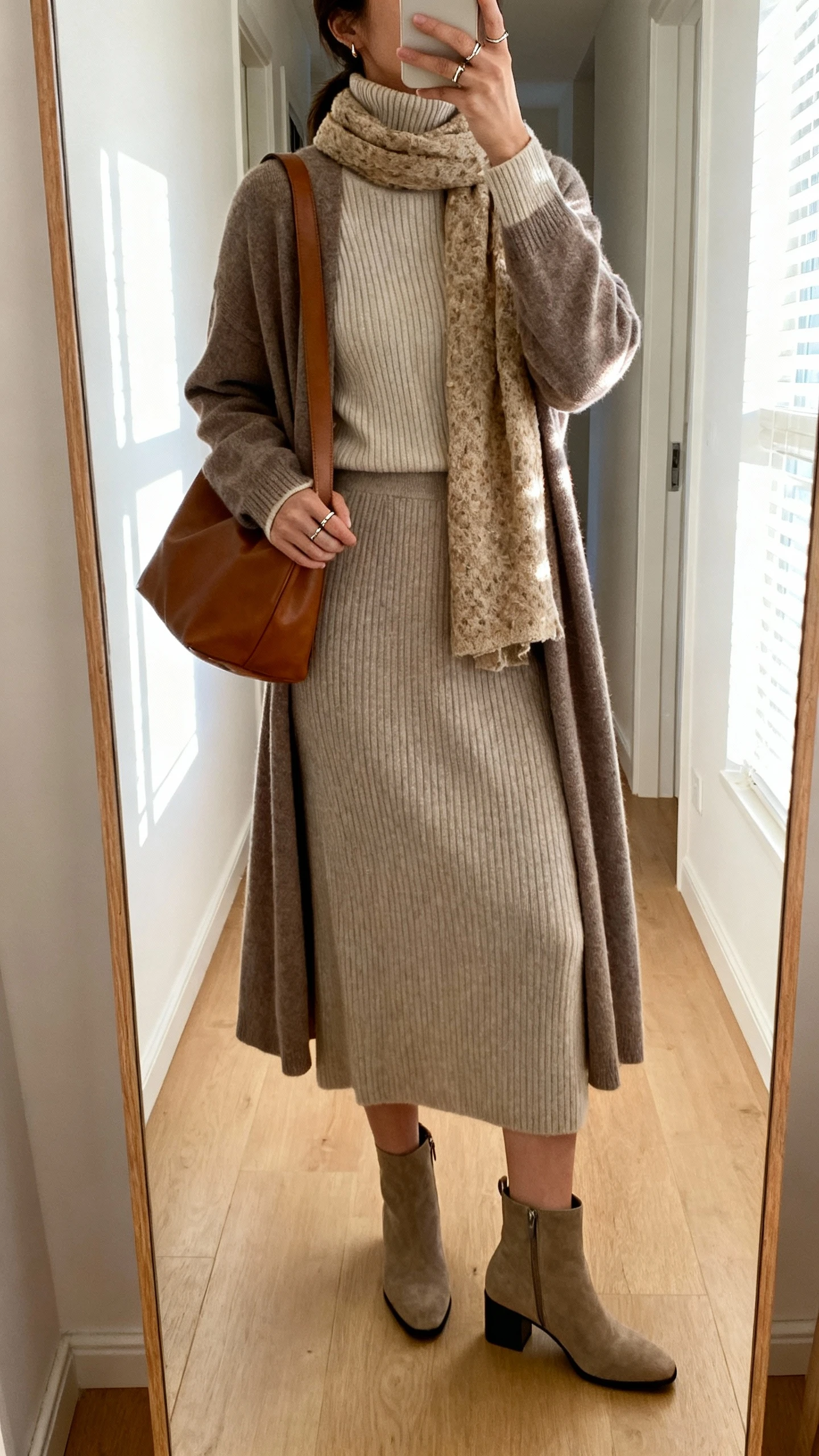 iPhone back-view mirror selfie of a woman wearing a fine-knit mock-neck sweater with a matching knit midi skirt, low block-heel ankle boots, a longline cardigan layered over, a textured scarf in a natural tone, a leather tote on shoulder, and minimal rings; face not visible, hallway full-length mirror, morning window light, iPhone photo quality.