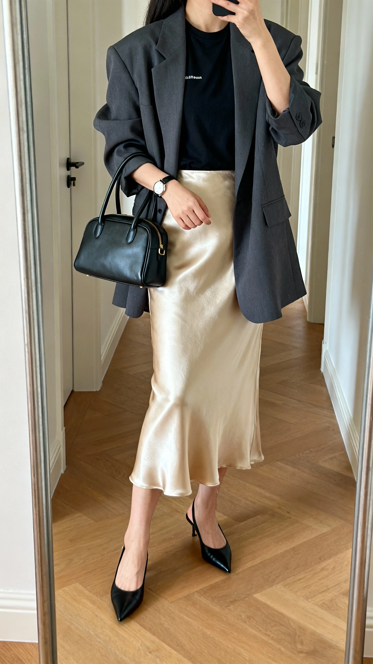 iPhone back-view mirror selfie of a woman wearing a champagne satin midi skirt, oversized charcoal blazer, fitted black tee, pointed black slingbacks, structured top-handle bag, and a sleek watch, face not visible, hallway mirror, natural daylight, iPhone photo quality.