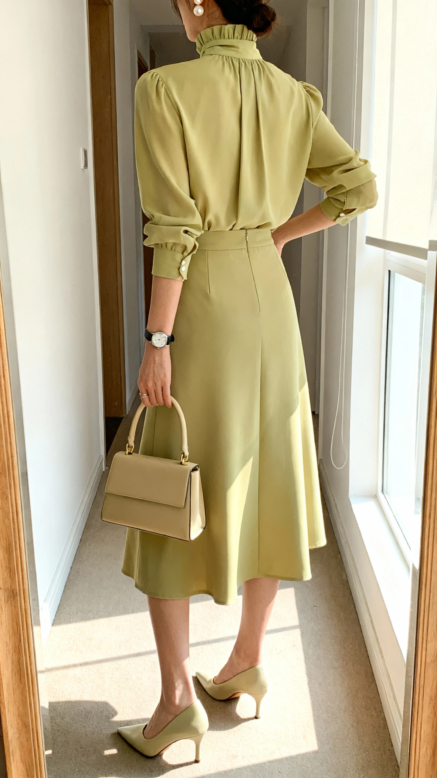 iPhone back-view mirror shot of a woman in a coordinated set: high-neck blouse with a matching midi skirt, pointed-toe pumps, structured top-handle bag, and pearl studs with a sleek watch; face not visible, full-length mirror in hallway, bright window light, iPhone photo quality.