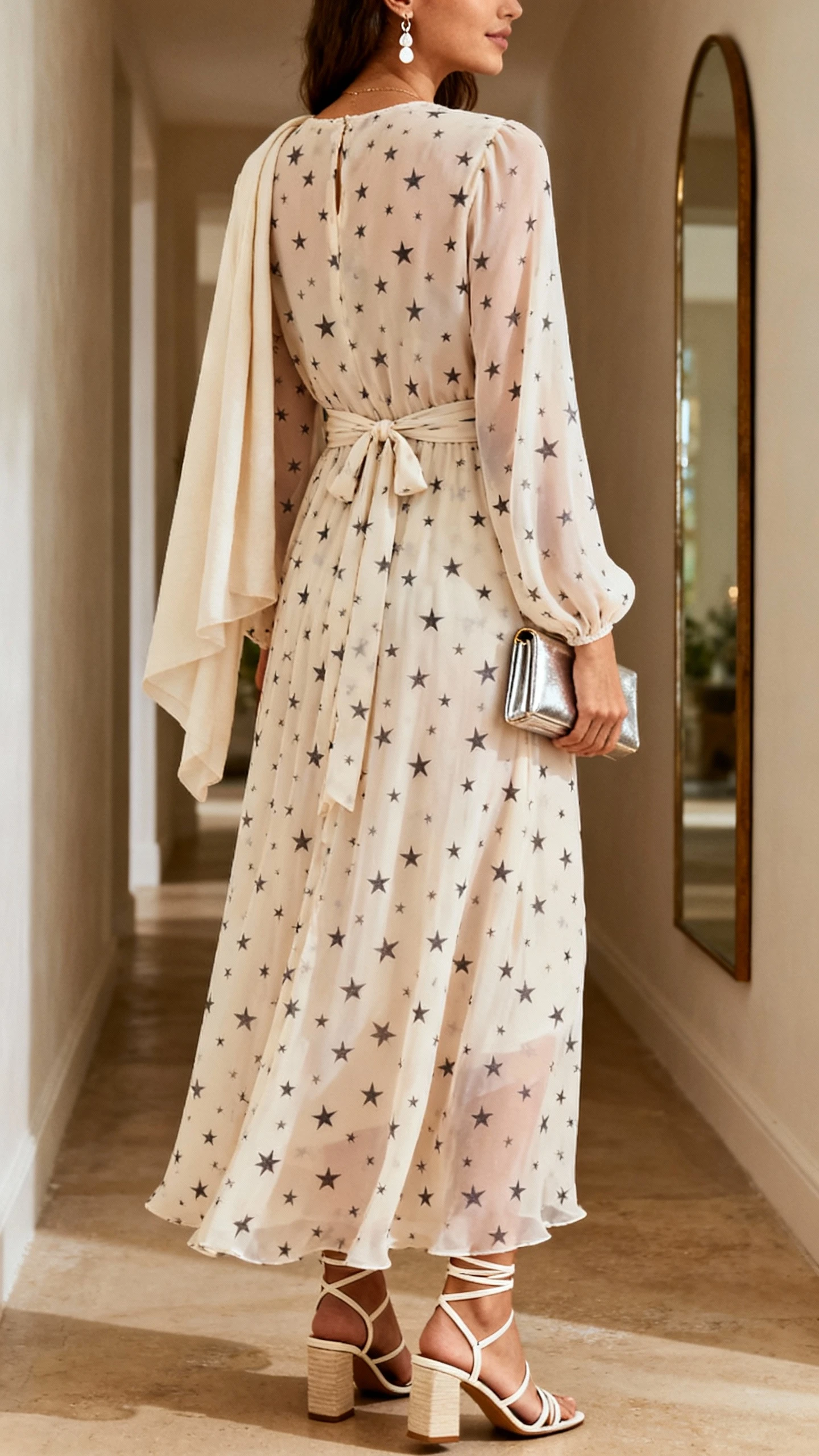 iPhone back view of a woman in a chiffon maxi dress with a micro-star print, wrap-front waist, strappy block-heel sandals, delicate drop earrings, a slim metallic clutch, and a lightweight shawl draped over shoulders; face not visible, hallway mirror, warm indoor lighting, iPhone photo quality.
