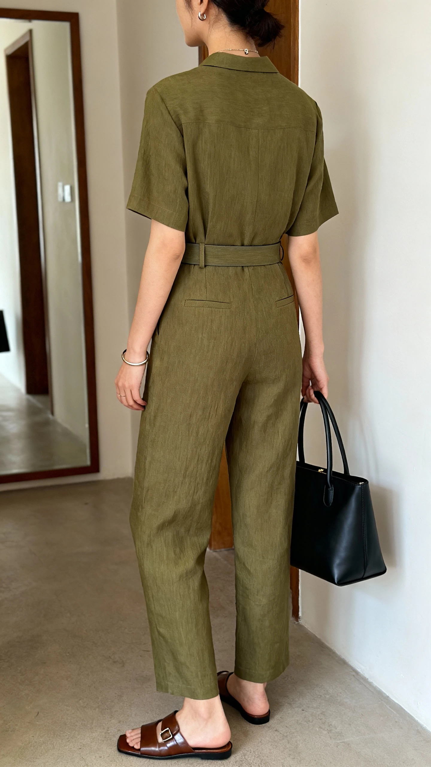 iPhone back view of a woman in a tailored linen jumpsuit in olive with polished leather slides, a simple bangle and small pendant earrings, a slim waist belt, and a medium top-handle bag in black, face not visible, hallway mirror, natural daylight, casual iPhone photo.