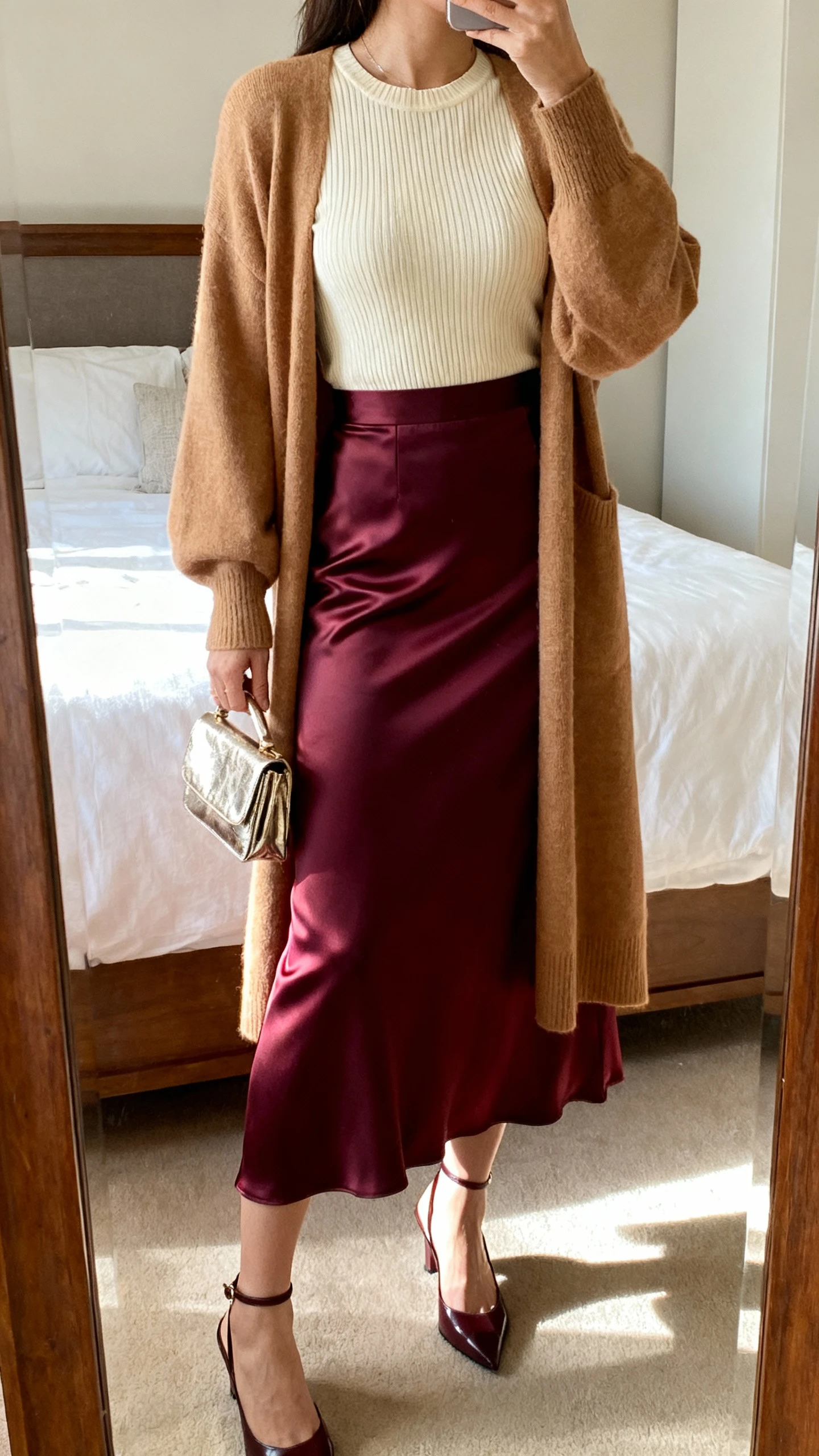 iPhone back view of a woman wearing a bias-cut satin maxi skirt in deep berry, a longline tan cardigan, a fitted cream ribbed knit top tucked in, slingback heels, and a small metallic clutch; face not visible, bedroom mirror, warm indoor lighting, iPhone photo quality.