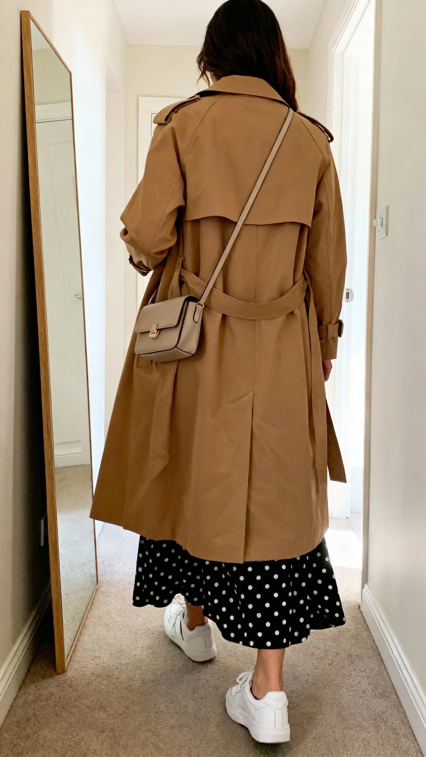 iPhone back view of a woman wearing a black polka dot maxi dress with a camel trench coat and clean white sneakers, small crossbody bag, face not visible, hallway full-length mirror, indoor daylight, iPhone photo quality.