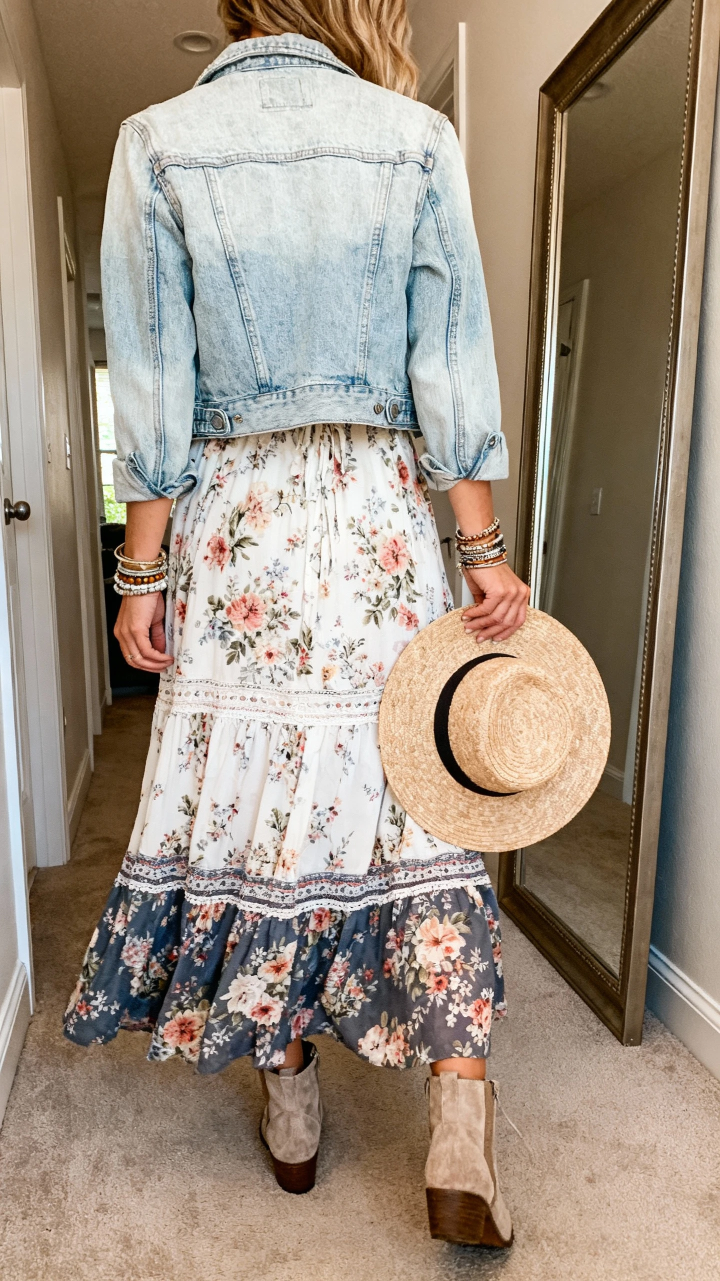iPhone back view of a woman wearing a breezy boho floral maxi dress layered with a light wash denim jacket, ankle boots, layered bracelets, and a wide-brim straw hat in hand, face not visible, hallway full-length mirror, indoor daylight, casual iPhone photo.