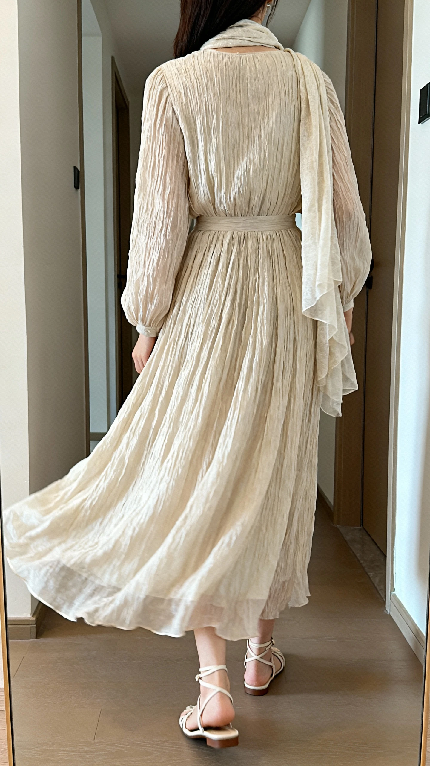 iPhone back view of a woman wearing a crinkle chiffon maxi dress with gentle pleats, long sleeves, and a flowing A-line skirt, styled with delicate strap flats and a lightweight shawl; face not visible, hallway full-length mirror, soft indoor daylight, casual iPhone photo.