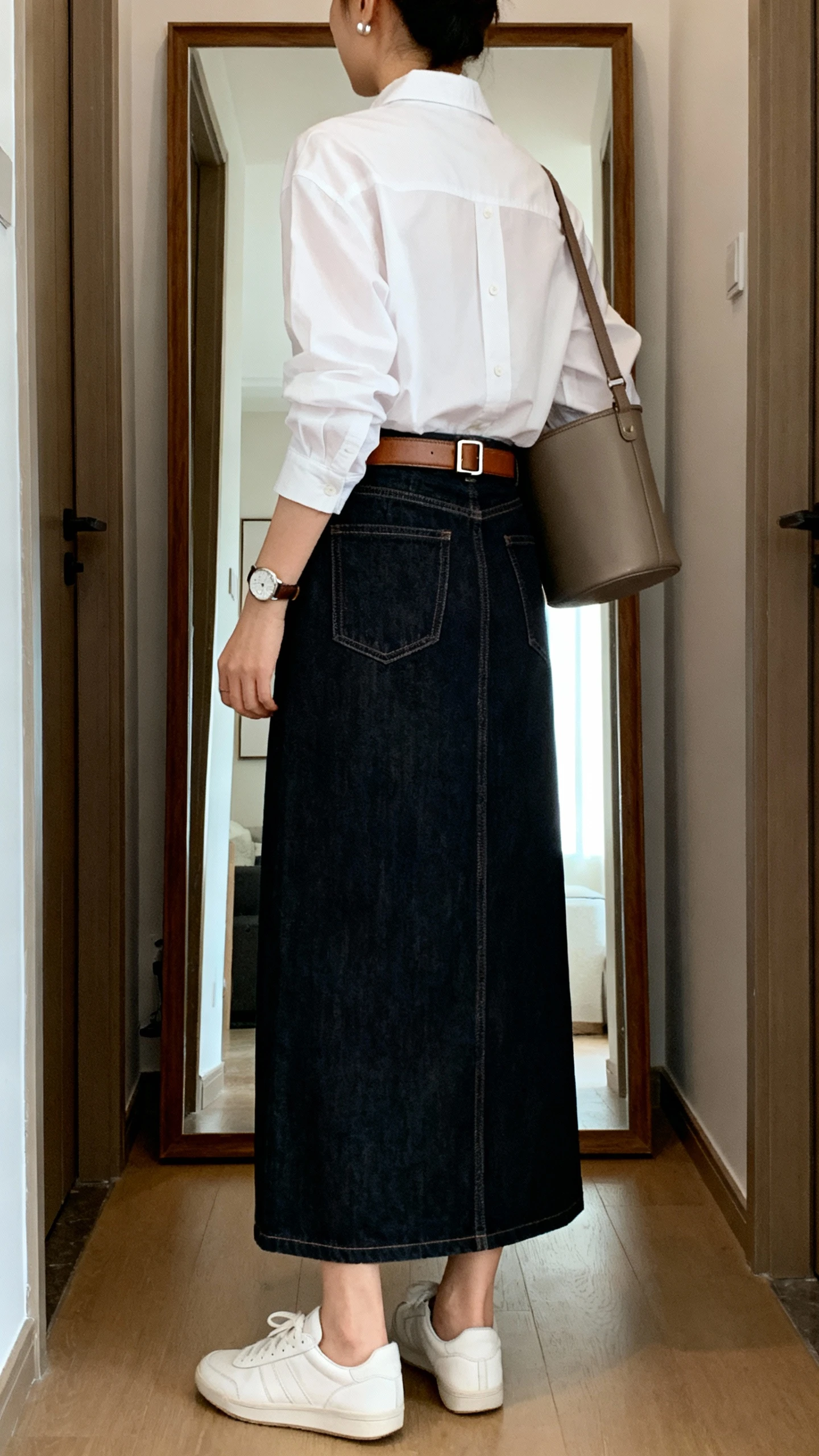 iPhone back view of a woman wearing a dark-wash denim maxi skirt with a straight cut, a crisp white button-down shirt tucked in, a leather belt with a subtle buckle, low-profile white sneakers, a classic watch, understated stud earrings, and a bucket bag on shoulder, face not visible, hallway full-length mirror, indoor daylight, iPhone photo quality.