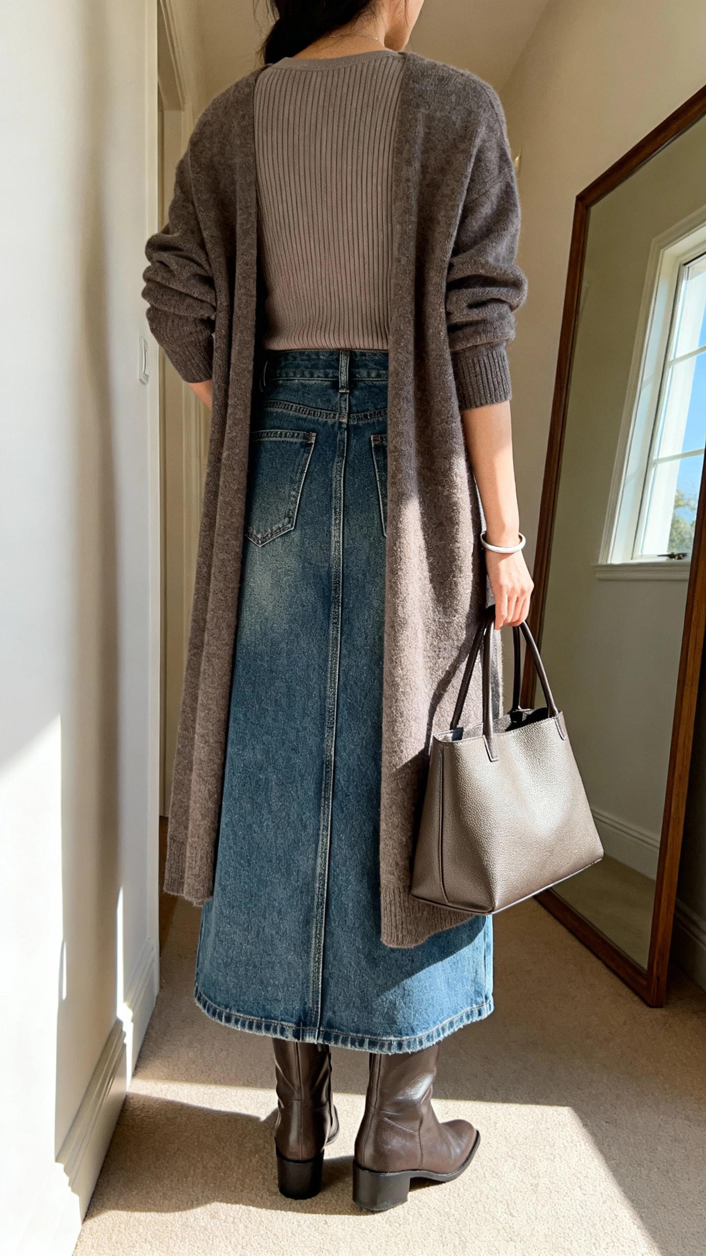 iPhone back view of a woman wearing a denim maxi skirt and a longline cardigan layered over a fitted ribbed top, ankle boots, structured tote, simple bracelet, face not visible, hallway mirror, indoor daylight from window, iPhone photo quality.