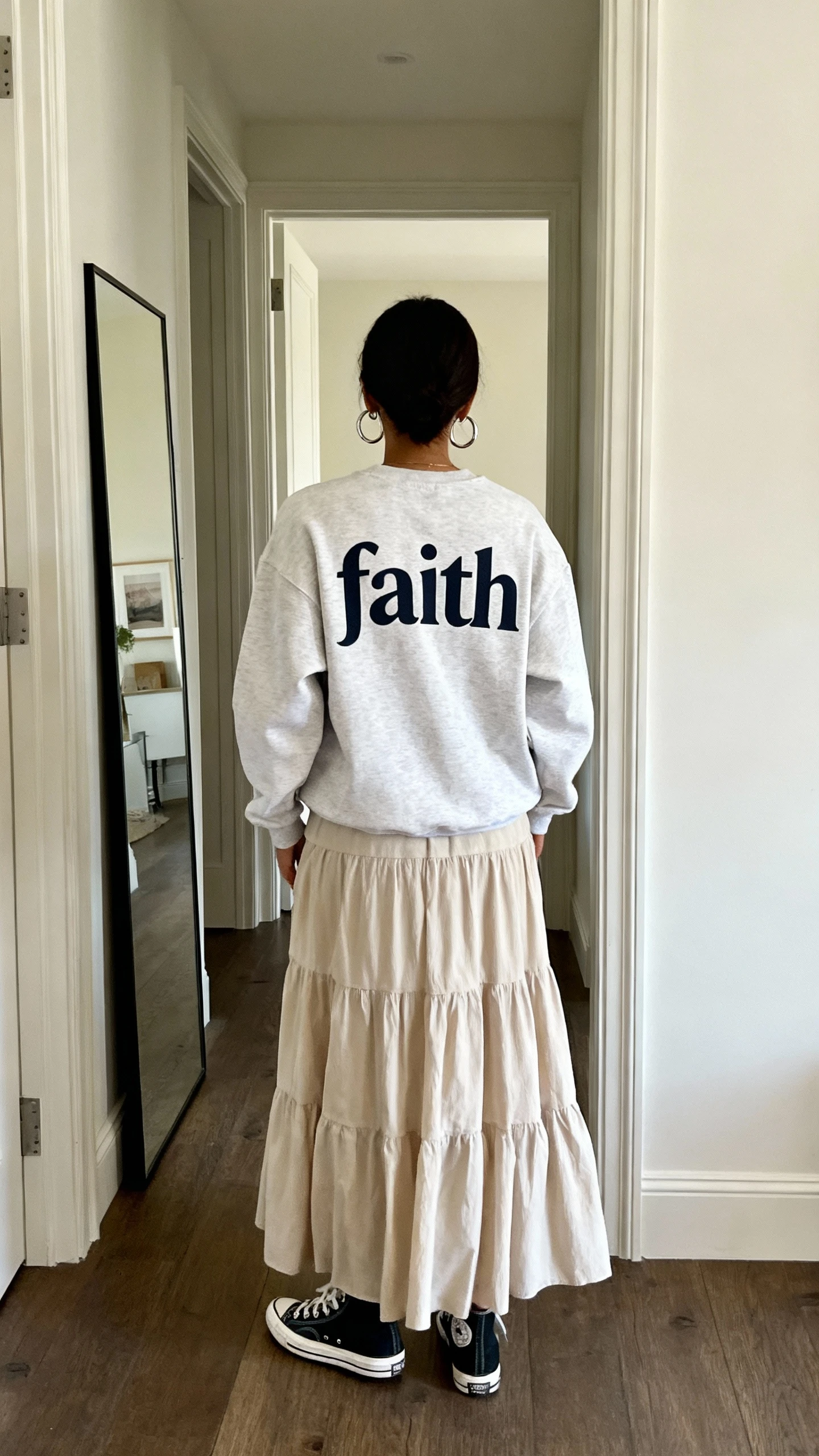 iPhone back view of a woman wearing a faith graphic crewneck with a tiered maxi skirt, styled with hoop earrings and low-top canvas sneakers, face not visible, hallway full-length mirror, natural daylight, casual iPhone photo