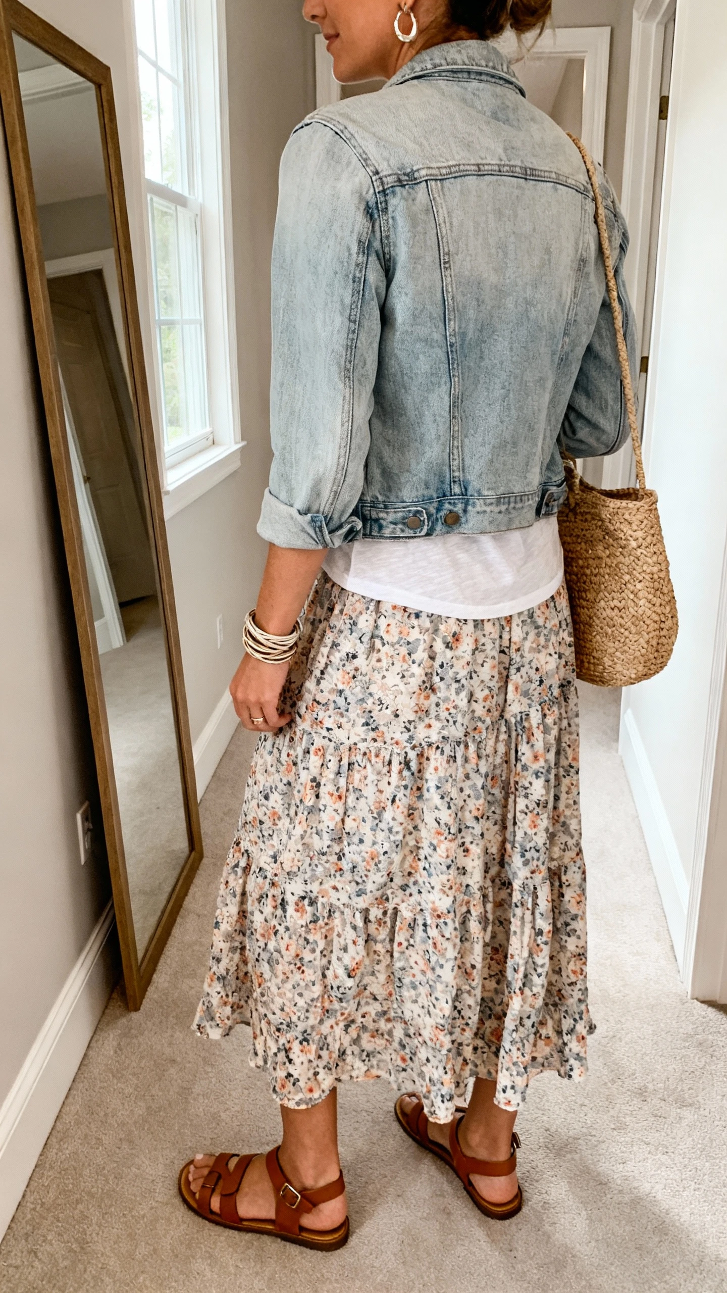 iPhone back view of a woman wearing a floral flowy maxi skirt, soft cotton tee, classic light-wash denim jacket, leather sandals, subtle drop earrings, wrap bracelet, and a woven straw bag; face not visible, hallway full-length mirror, morning window light, casual iPhone photo.