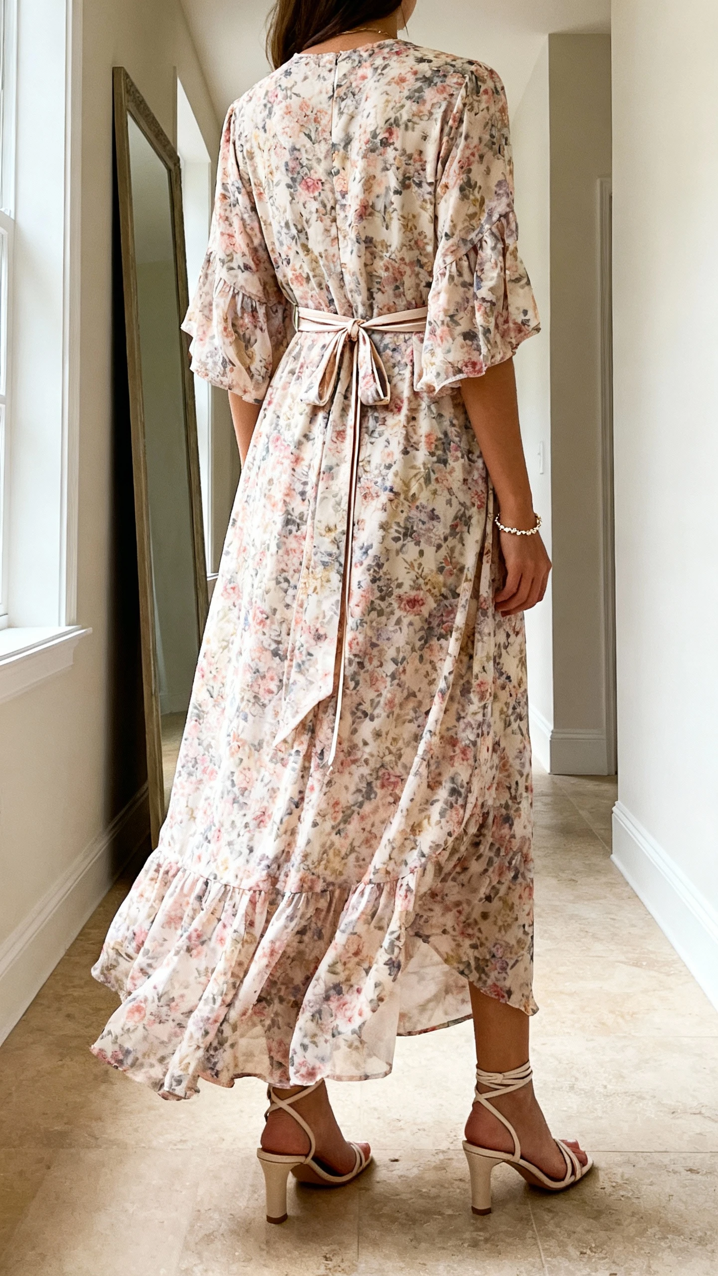 iPhone back view of a woman wearing a flowing floral maxi dress with an elegant wrap detail, thin waist tie, dainty bracelet, and strappy heeled sandals, face not visible, hallway mirror, natural daylight from window, iPhone photo quality.