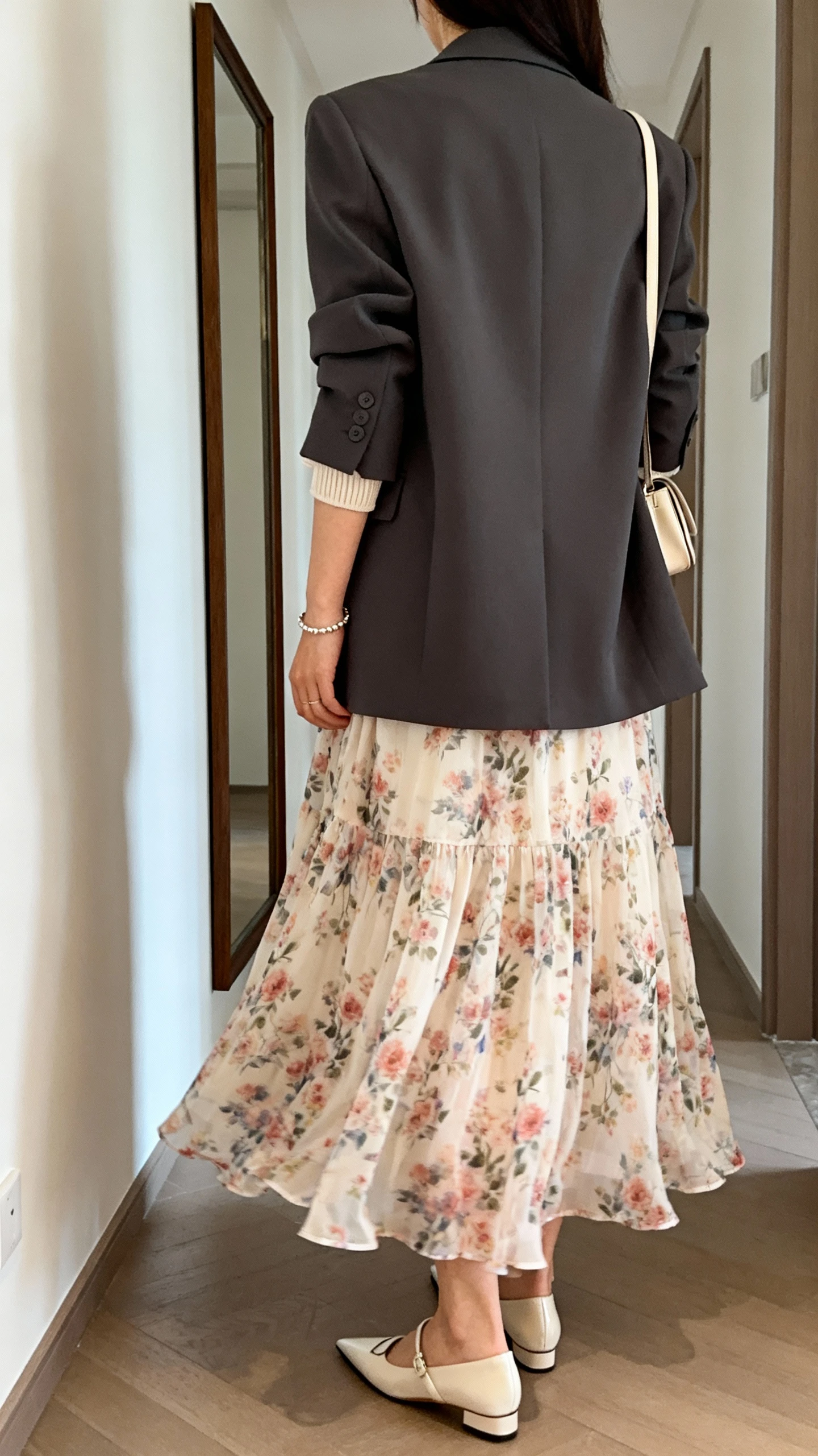 iPhone back view of a woman wearing a flowing floral midi skirt with a structured blazer, tucked simple shell top, dainty bracelet, pointed-toe flats, and a minimal shoulder bag, face not visible, hallway mirror, soft natural daylight, iPhone photo quality.