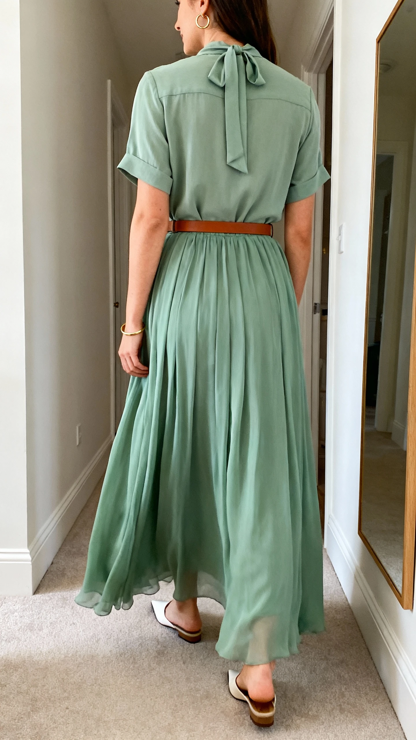 iPhone back view of a woman wearing a flowy sage chiffon maxi skirt, a short-sleeve blouse with a modest tie-neck, a cognac slim leather belt, pointed-toe flats, and delicate gold hoop earrings and a thin gold bracelet, face not visible, hallway mirror, soft morning light, casual iPhone photo.