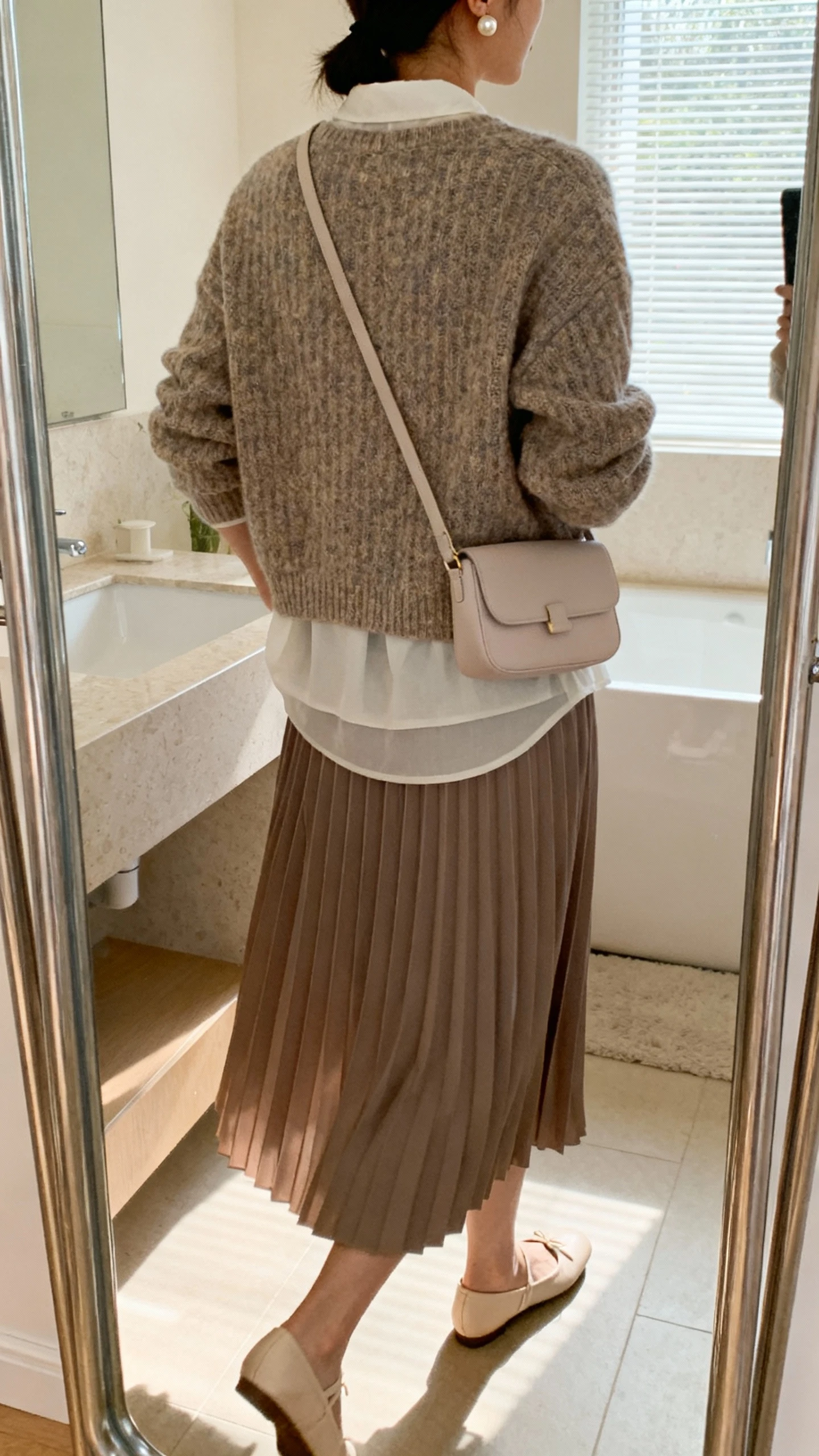 iPhone back view of a woman wearing a layered knit sweater over a pleated midi skirt with ballet flats, pearl stud earrings, and a soft crossbody bag, face not visible, gentle walking pose in a bathroom mirror, soft window light, iPhone photo quality.
