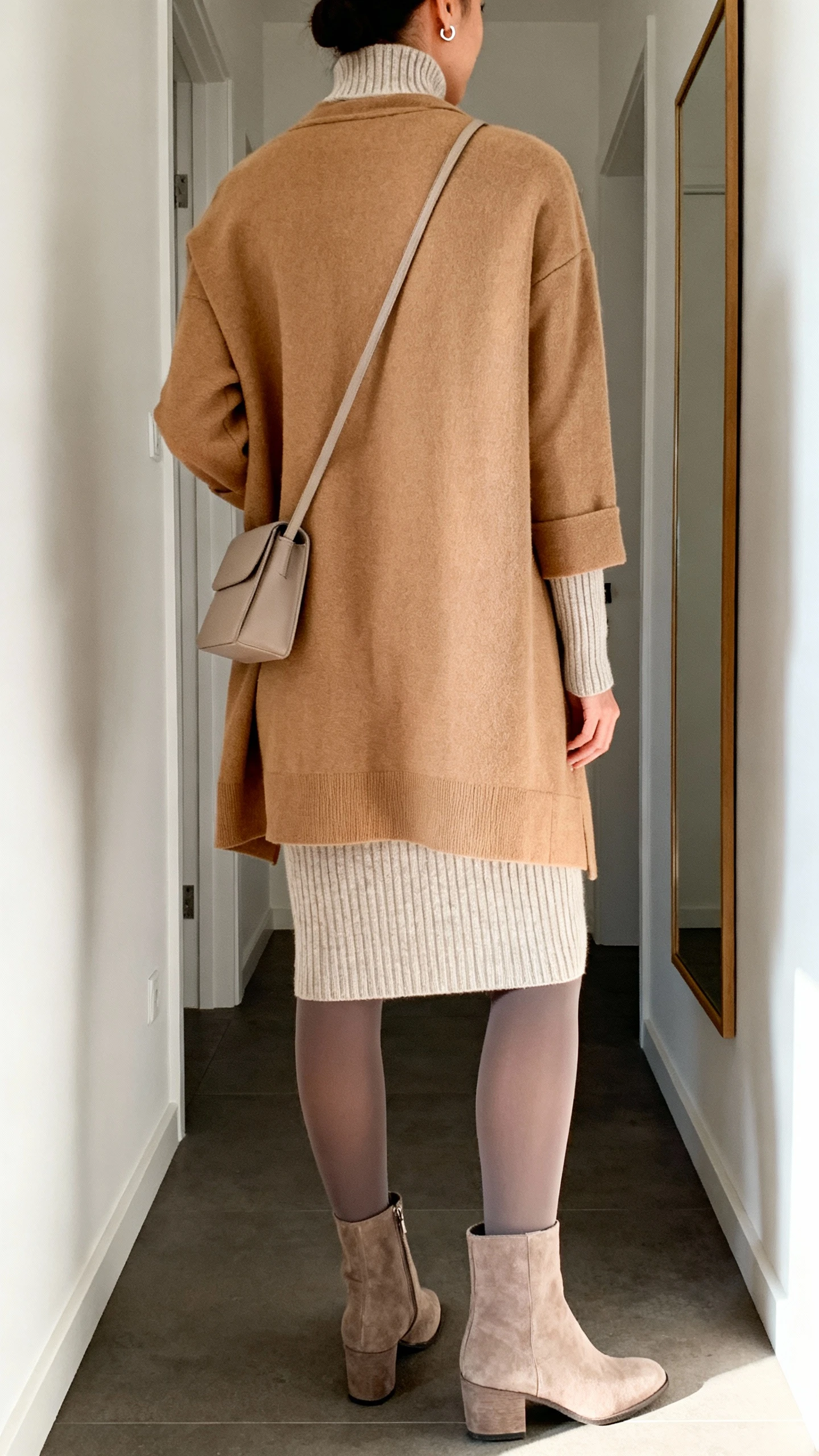 iPhone back view of a woman wearing a long-sleeve knit dress layered with a camel coatigan, opaque tights, ankle boots, simple stud earrings, and a minimal crossbody bag, face not visible, hallway mirror, morning indoor light, casual iPhone photo.