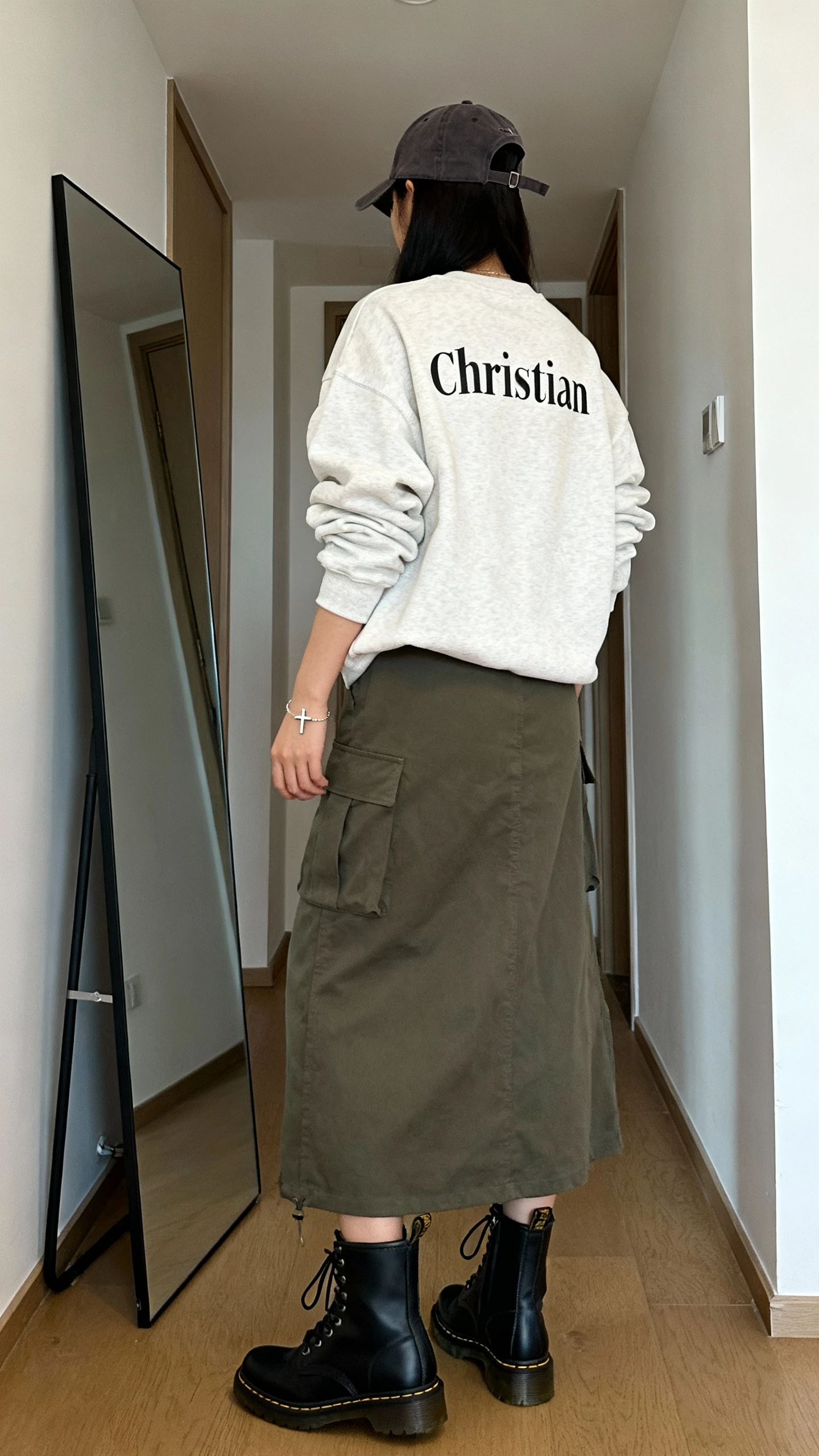 iPhone back view of a woman wearing a modest Christian crewneck with a cargo maxi skirt, lace-up ankle boots, baseball cap, and a minimal cross bracelet; face not visible, hallway full-length mirror, natural daylight, iPhone photo quality.
