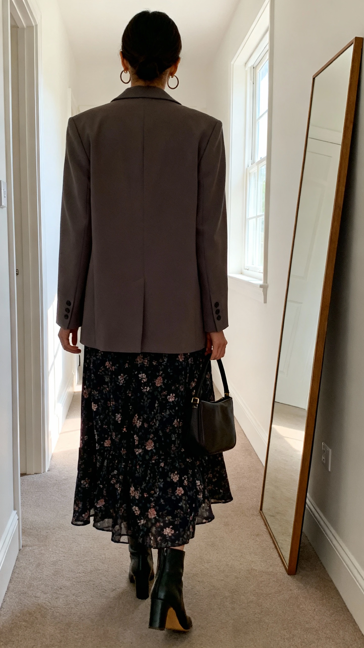 iPhone back view of a woman wearing a moody dark floral maxi dress with a soft tailored blazer, minimalist hoop earrings, structured handbag, and heeled ankle boots, face not visible, hallway full-length mirror, natural daylight from window, iPhone photo quality.